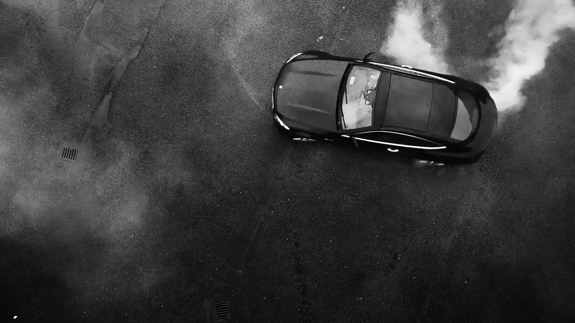 An overhead shot of a black car doing a burnout on a dark, wet asphalt surface, creating a cloud of smoke.