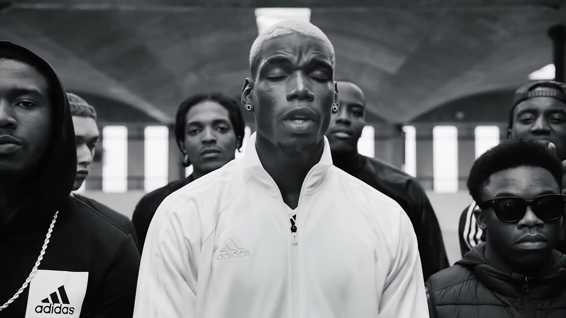 Paul Pogba, a Black man with bleached hair, stands in a white track jacket, surrounded by a diverse group of young men in a large, arched hall.