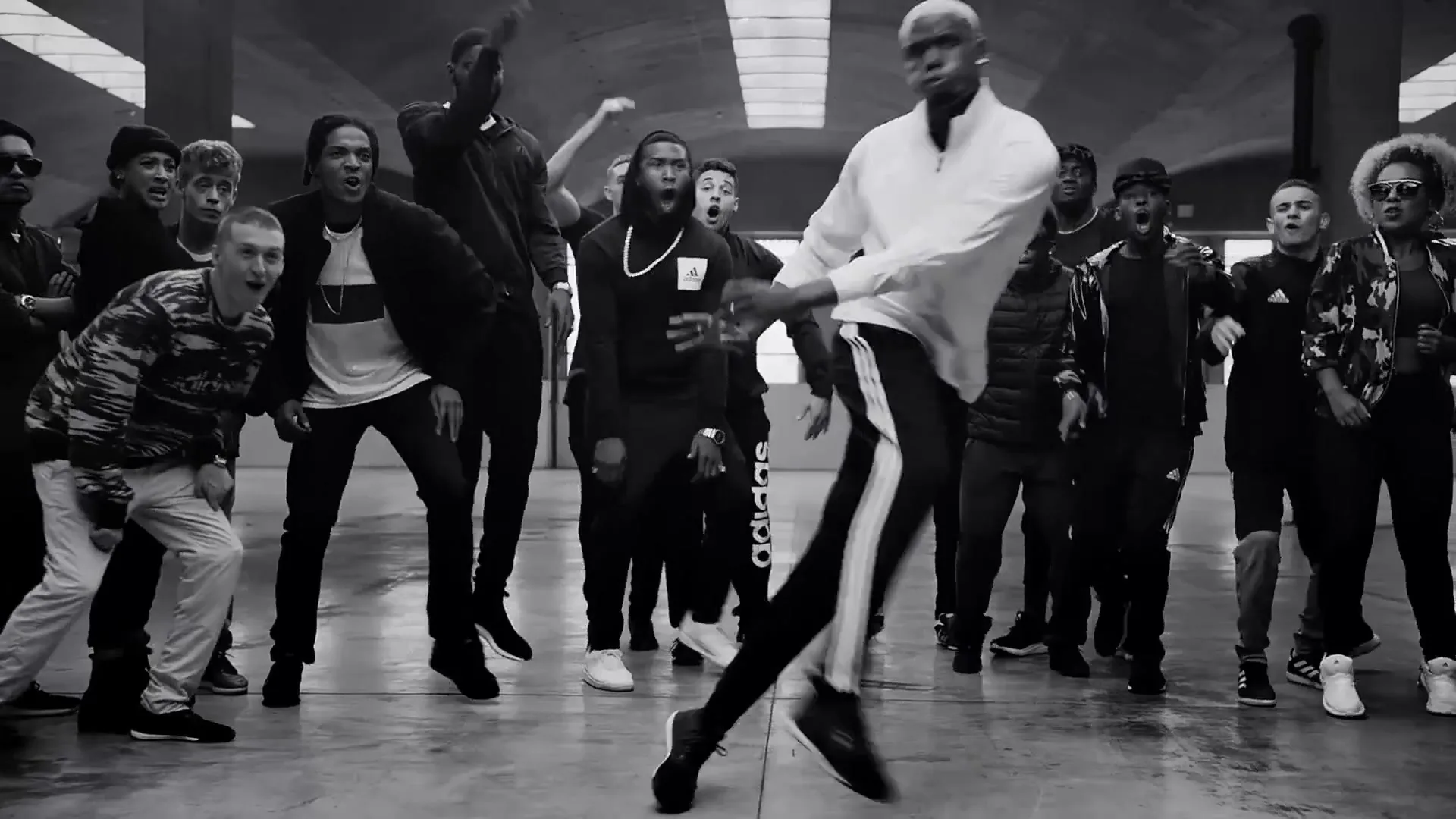 Paul Pogba, a male soccer player with bleached hair, dribbles a soccer ball while surrounded by a diverse group of people in an indoor concrete space.