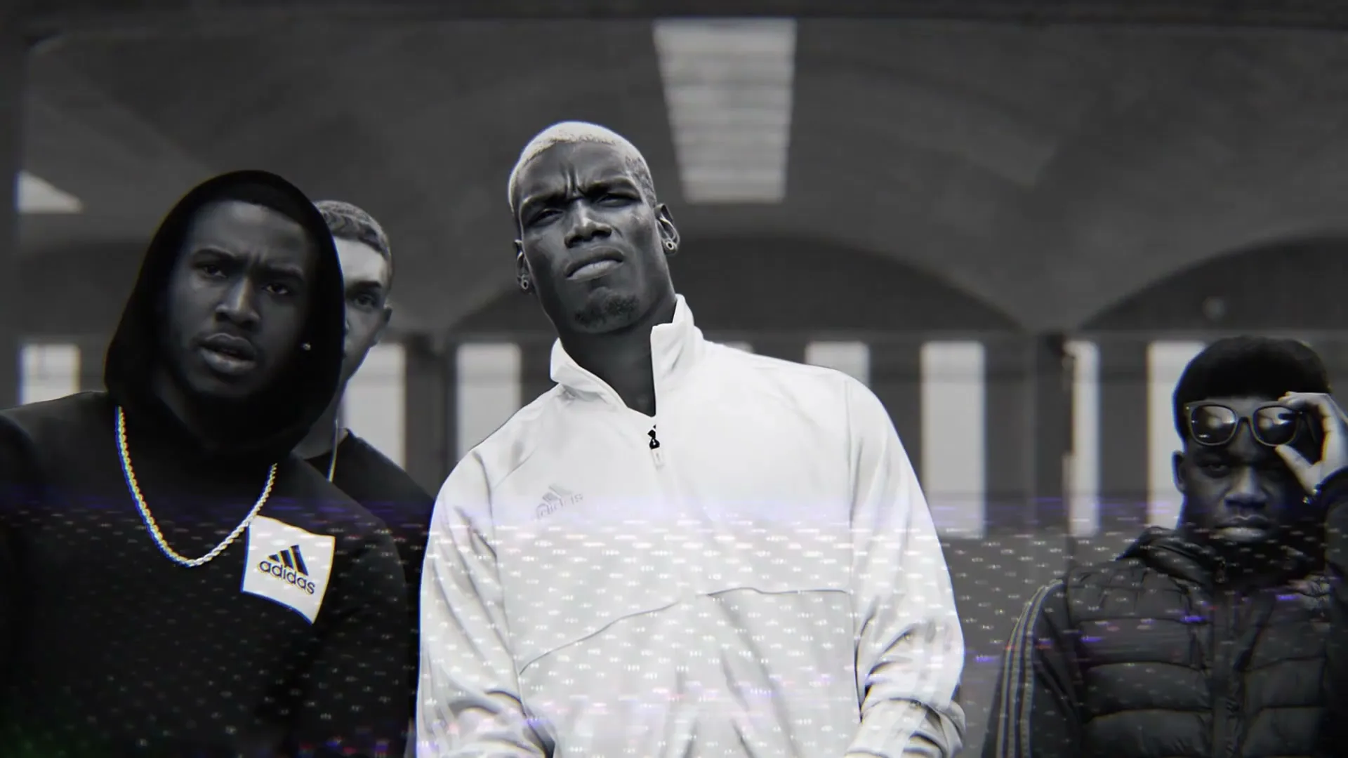 Paul Pogba, a male with bleached hair, stands in a black and white shot, surrounded by other men, looking directly at the camera with a serious expression.