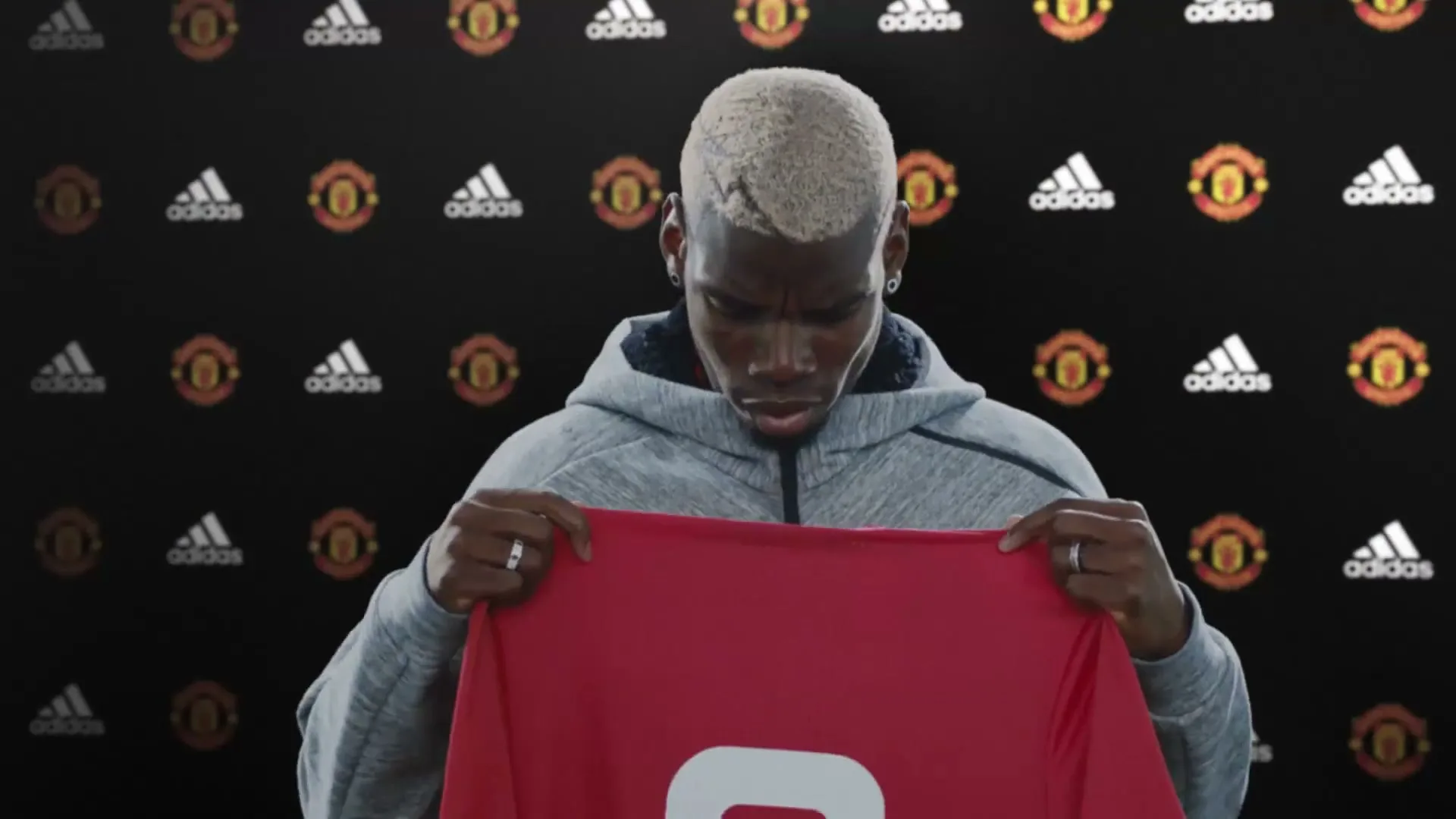 Paul Pogba, a male with bleached hair, holds up a red jersey with "POGBA" and the number "6" on it, in front of a black backdrop with adidas and Manchester United logos.