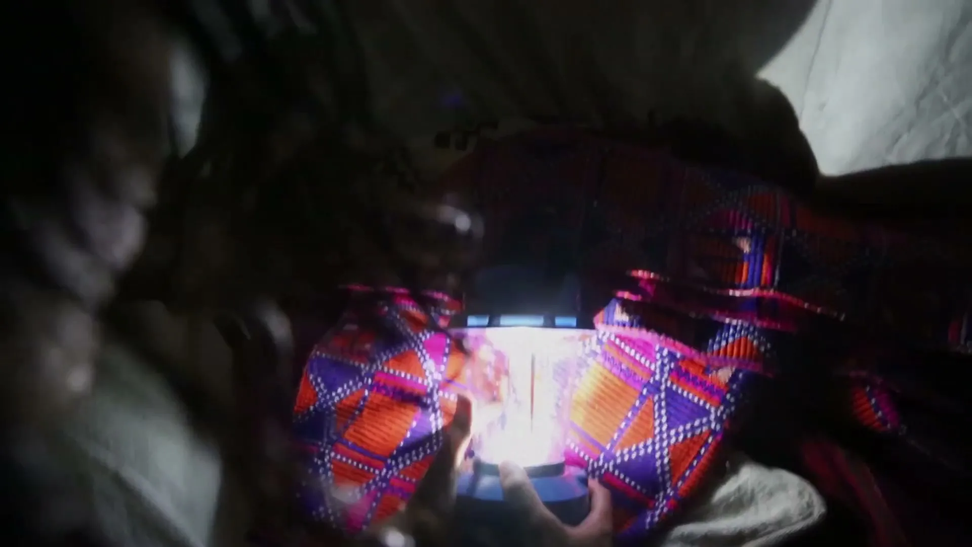 A person's hands hold a glowing lantern, illuminating a colorful patterned fabric in a dark setting.