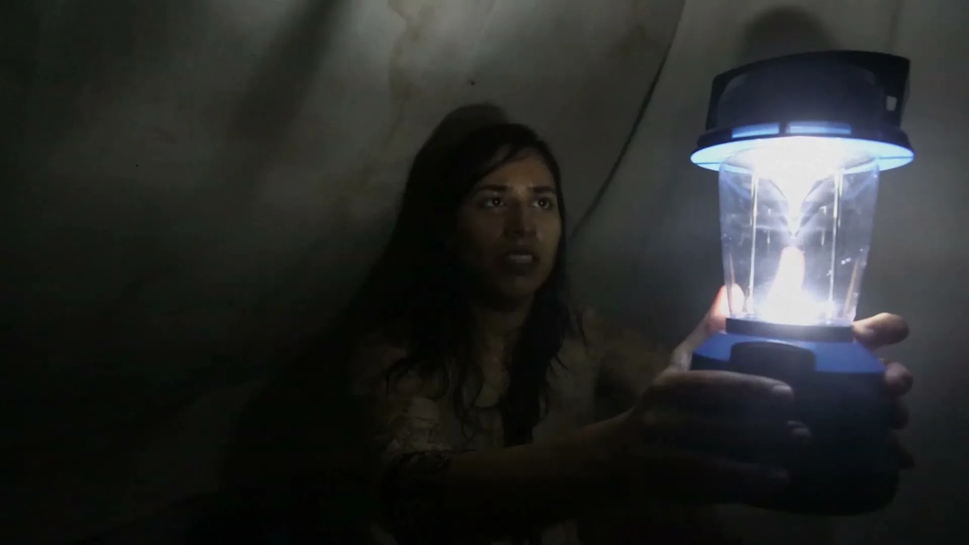A young woman with long dark hair, wearing a light-colored shirt, holds a bright lantern in a dark, confined space, looking around with a worried expression.