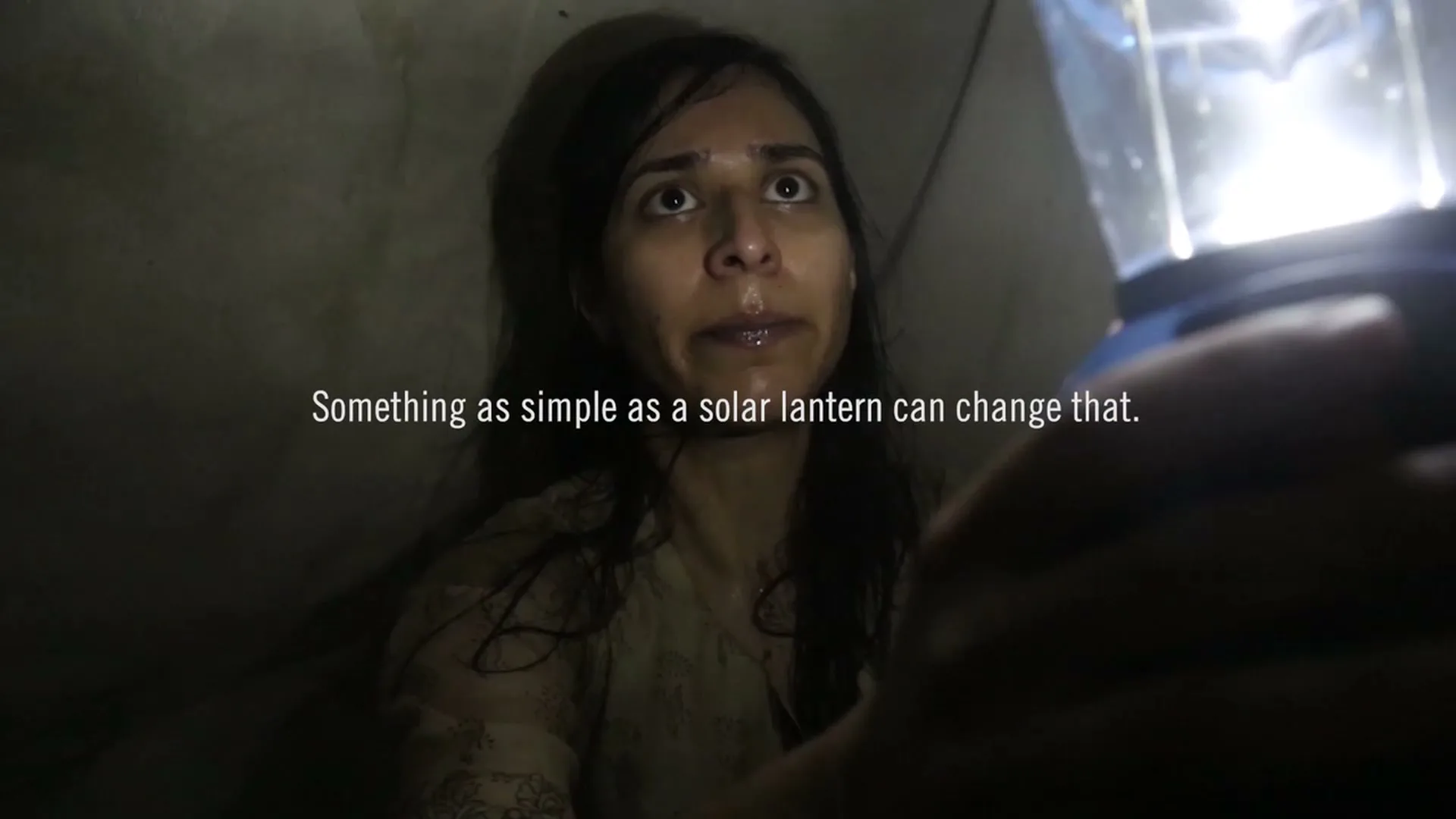 A young woman with dark hair and a light-colored shirt looks up with a worried expression as a bright solar lantern illuminates her face in a dark, confined space.
