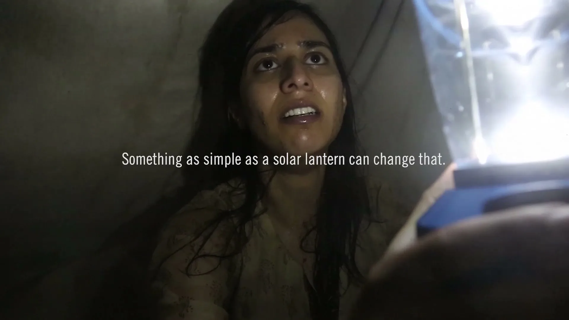 A young woman with wet hair and a distressed expression looks up at a solar lantern held by an unseen person in a dark, confined space.