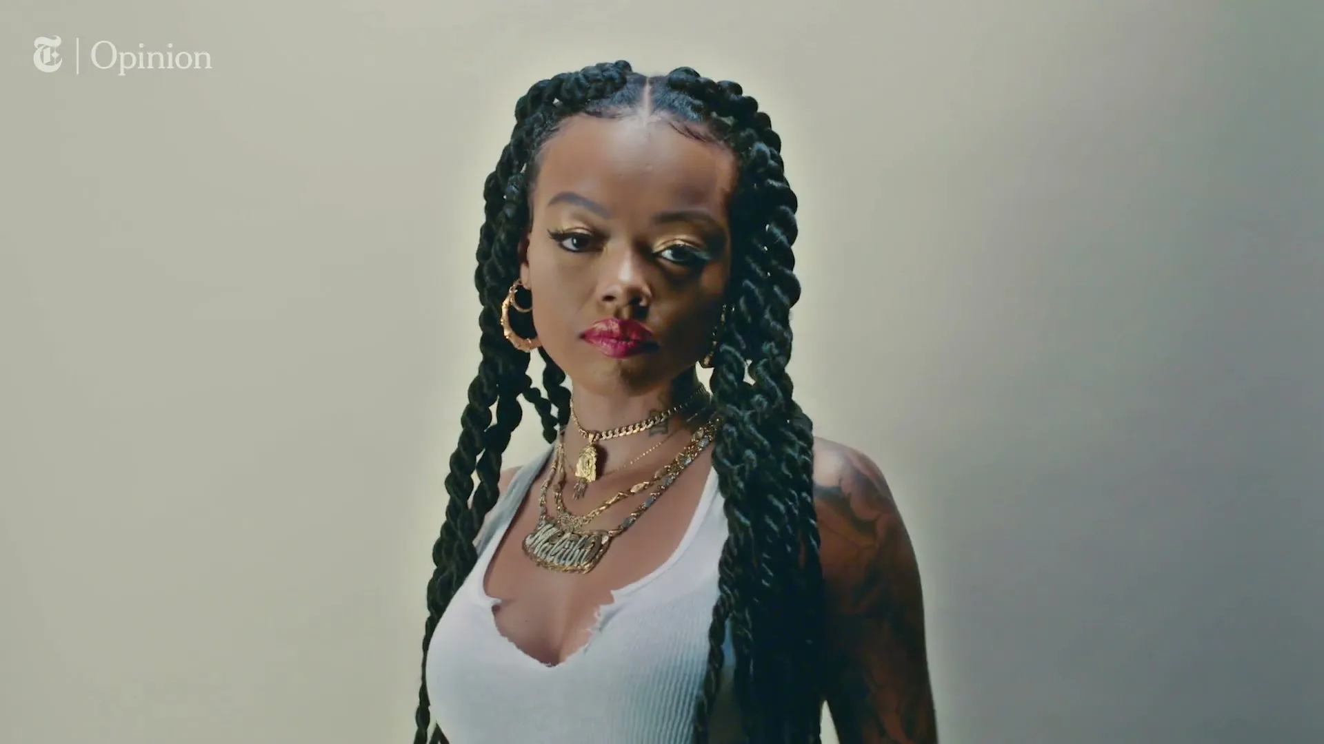 A young Black woman with long braids, wearing a white tank top and gold jewelry, looks directly at the camera in a studio.