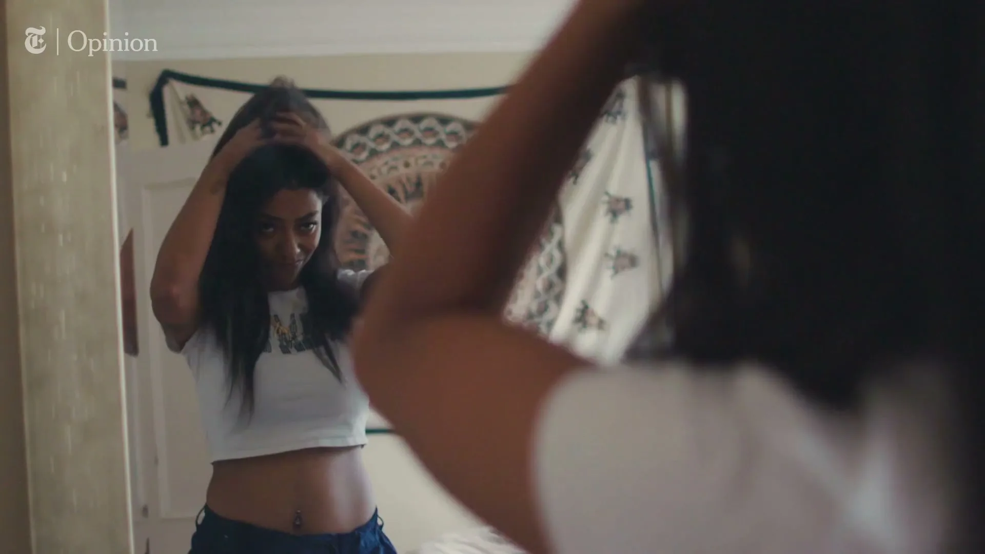 A young woman with long dark hair, wearing a white crop top, looks at her reflection in a mirror, adjusting her hair in a bedroom.