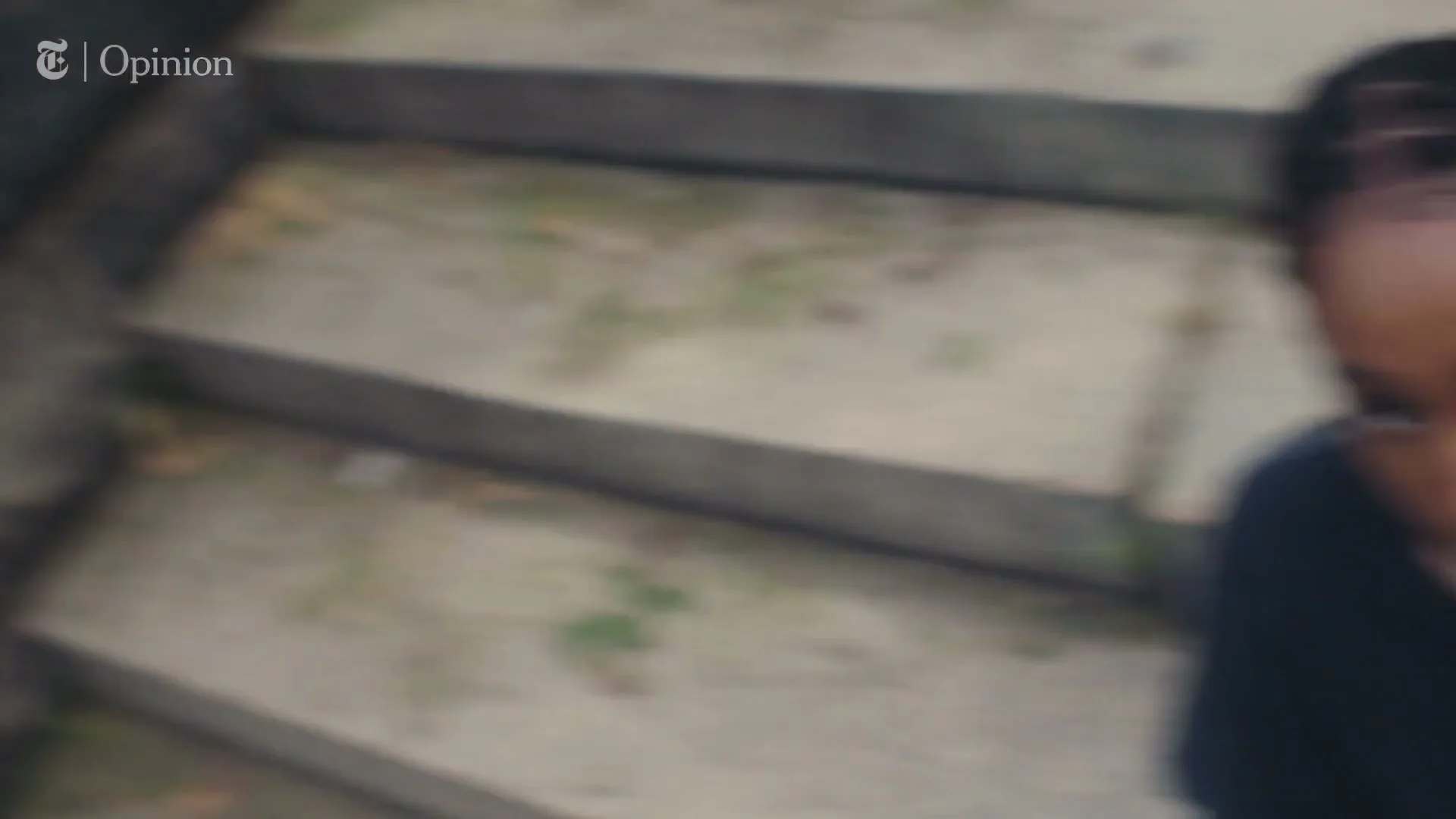 A young Black woman with braided hair and a black t-shirt looks up at the camera while sitting on concrete steps outdoors.