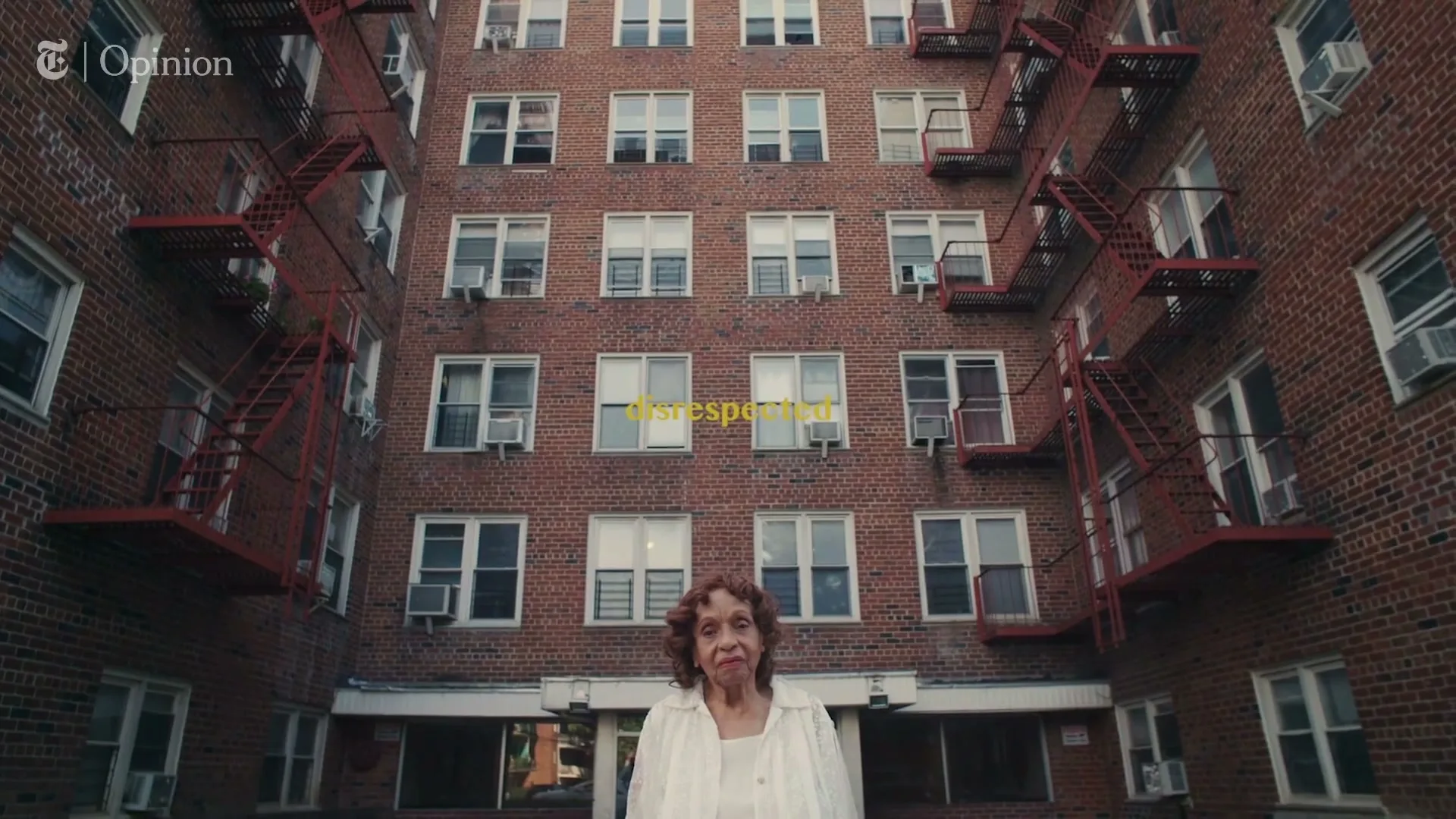 An older woman with curly brown hair and a white top stands in front of a large brick apartment building with many windows and red fire escapes.