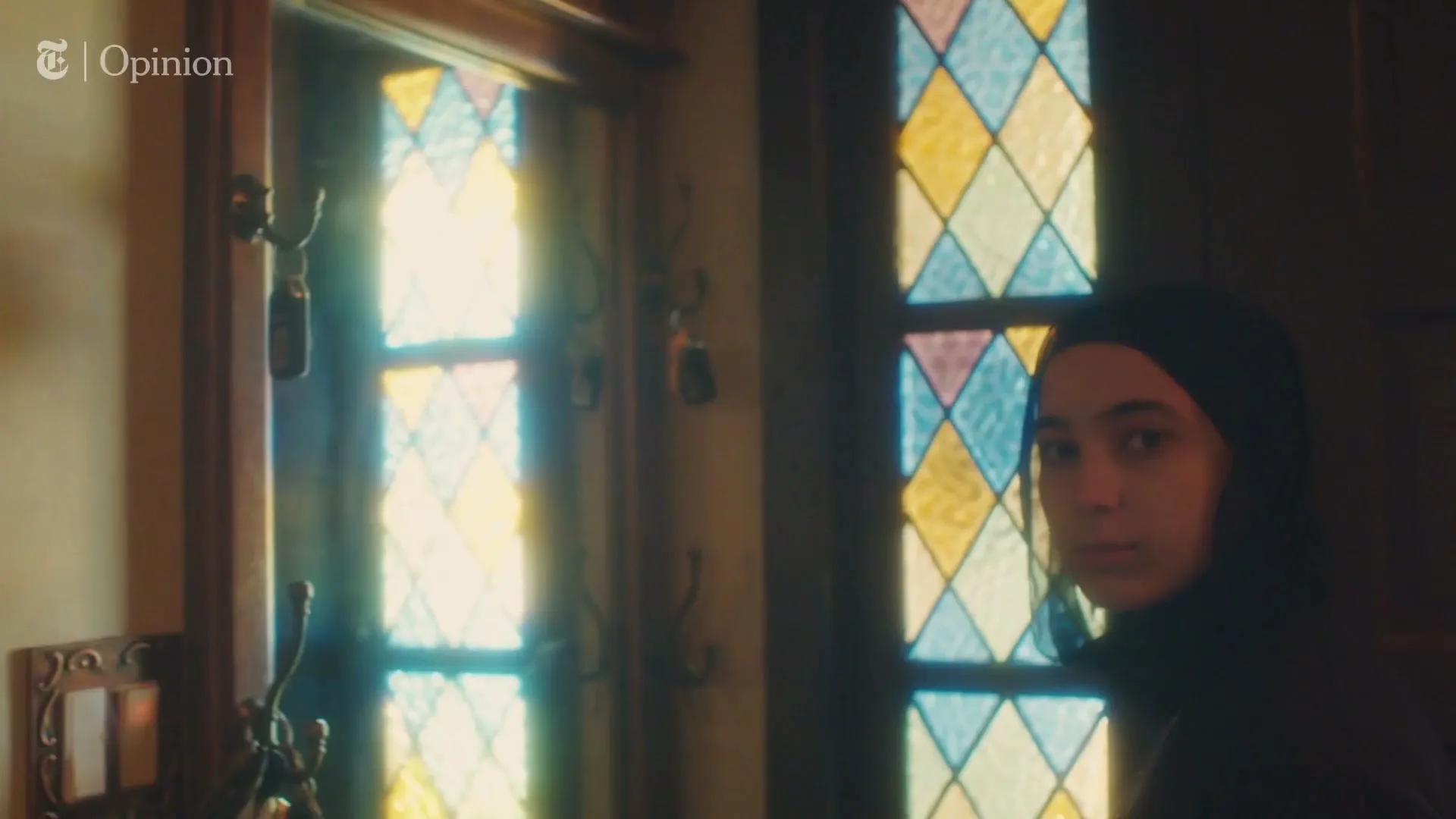 A young woman in a black hijab looks towards the camera, standing next to a stained glass window in a dimly lit room.