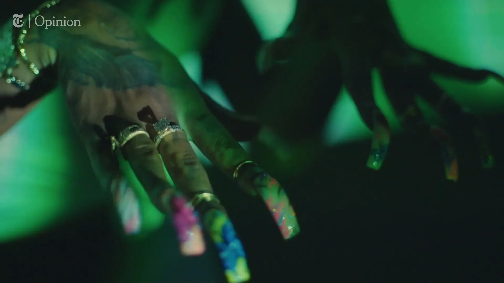 A person's hands with long, colorful nails and rings are shown in a dark, green-lit setting, with a "T | Opinion" logo in the corner.