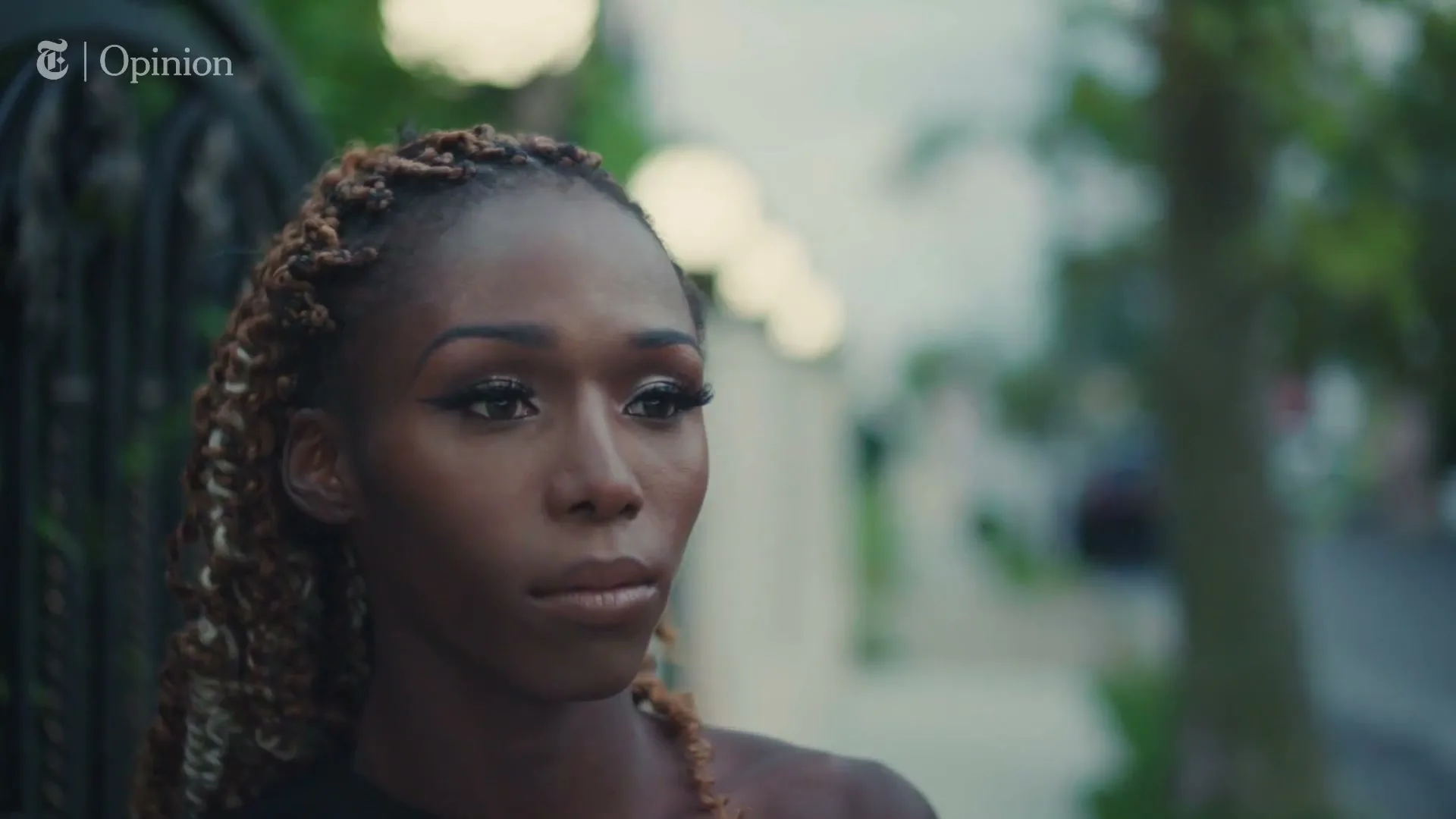 A young Black woman with braided hair looks off-camera with a serious expression, standing outdoors near a fence.