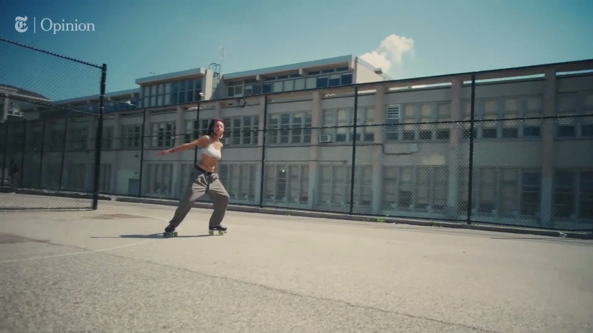 A young woman with long dark hair, wearing a white crop top and grey pants, roller skates on an outdoor court in front of a school building on a sunny day.