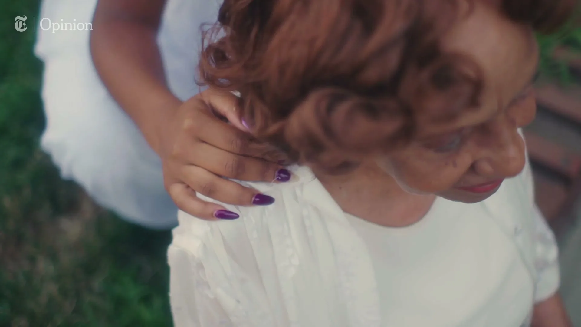 A young woman's hand with purple nails rests on the shoulder of an older woman with red hair, both wearing white, outdoors.