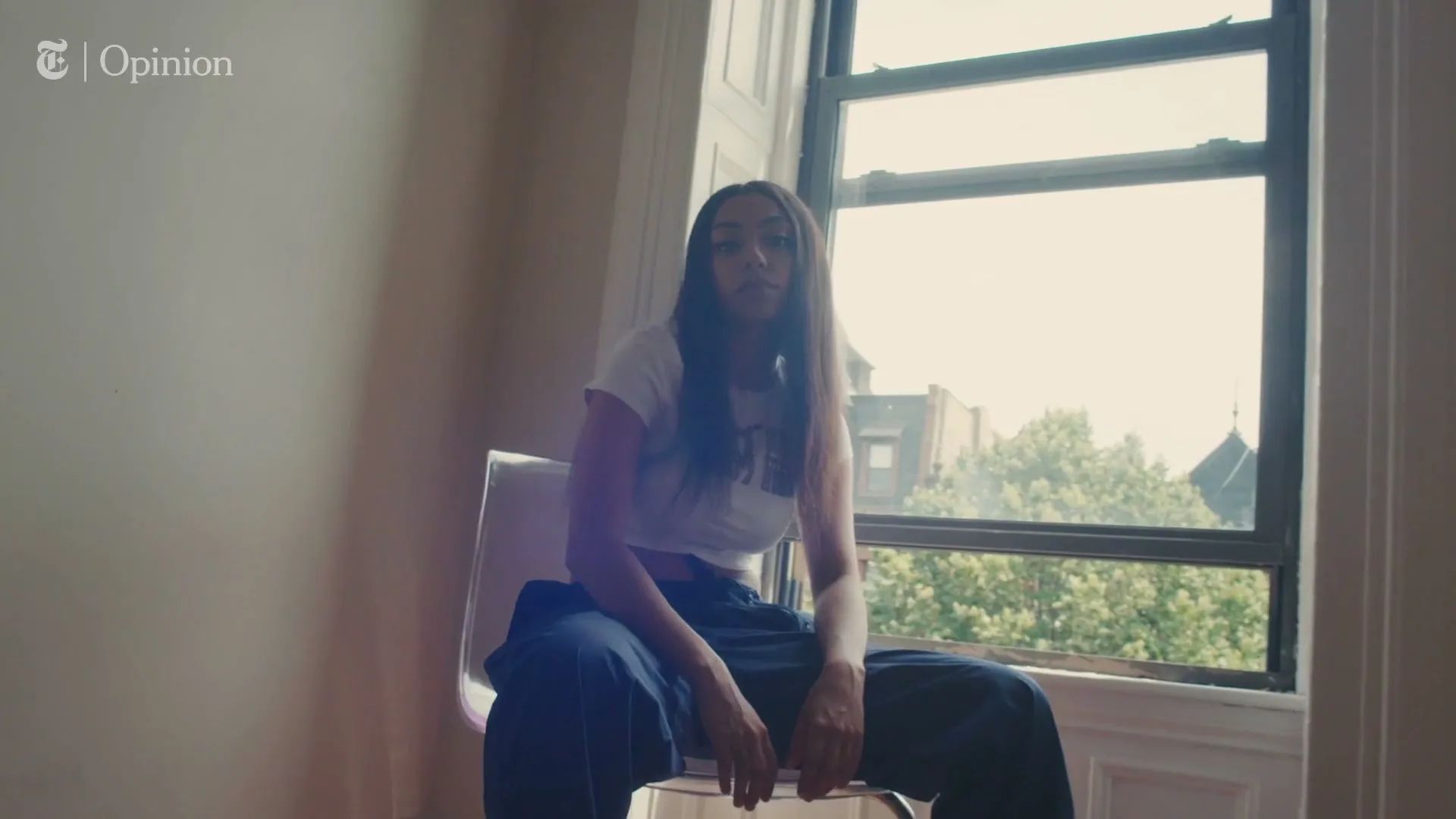 A young woman with long dark hair sits on a clear chair in front of a window, looking directly at the camera in a room with light-colored walls.