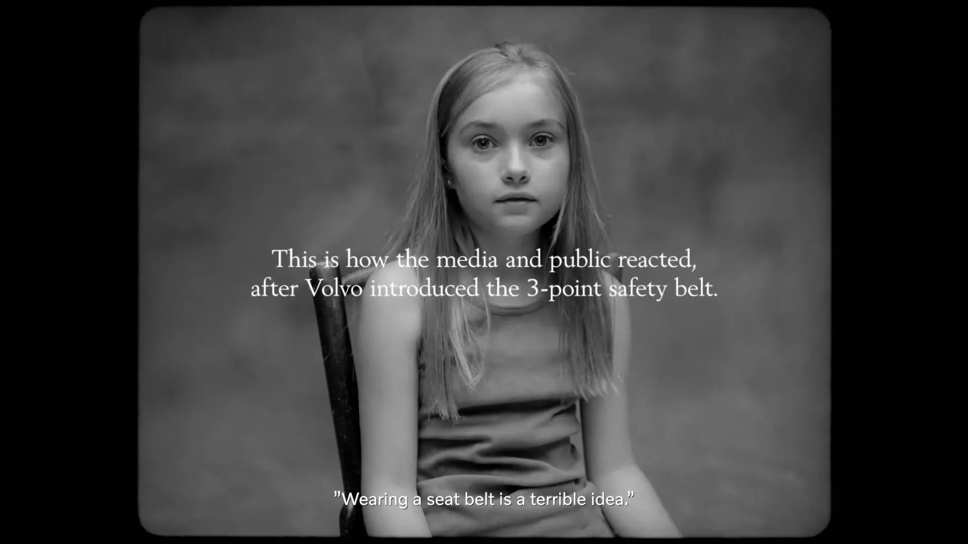 A young girl with long hair sits on a chair, looking down and then up at the camera, in a black and white studio setting.