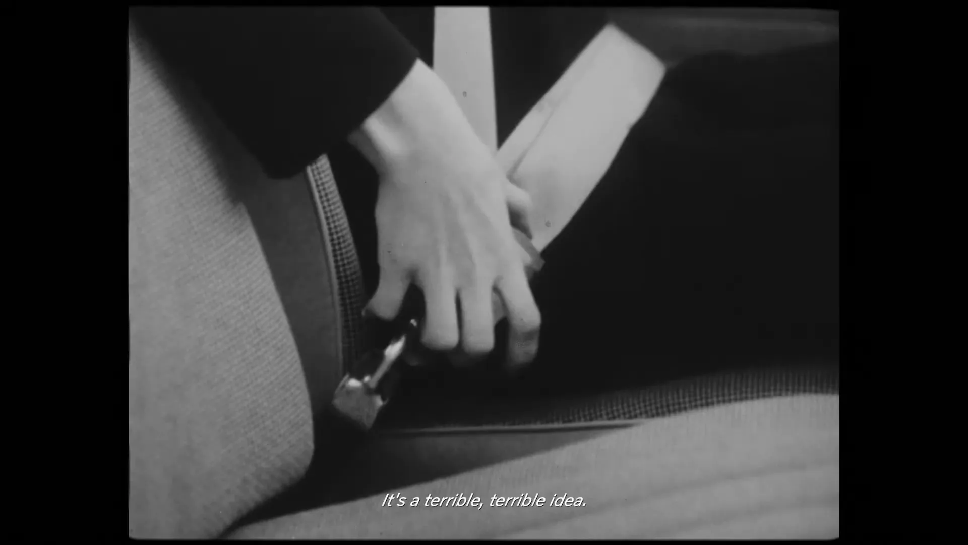 A person's hand unbuckles a seatbelt in a car, shown in black and white.