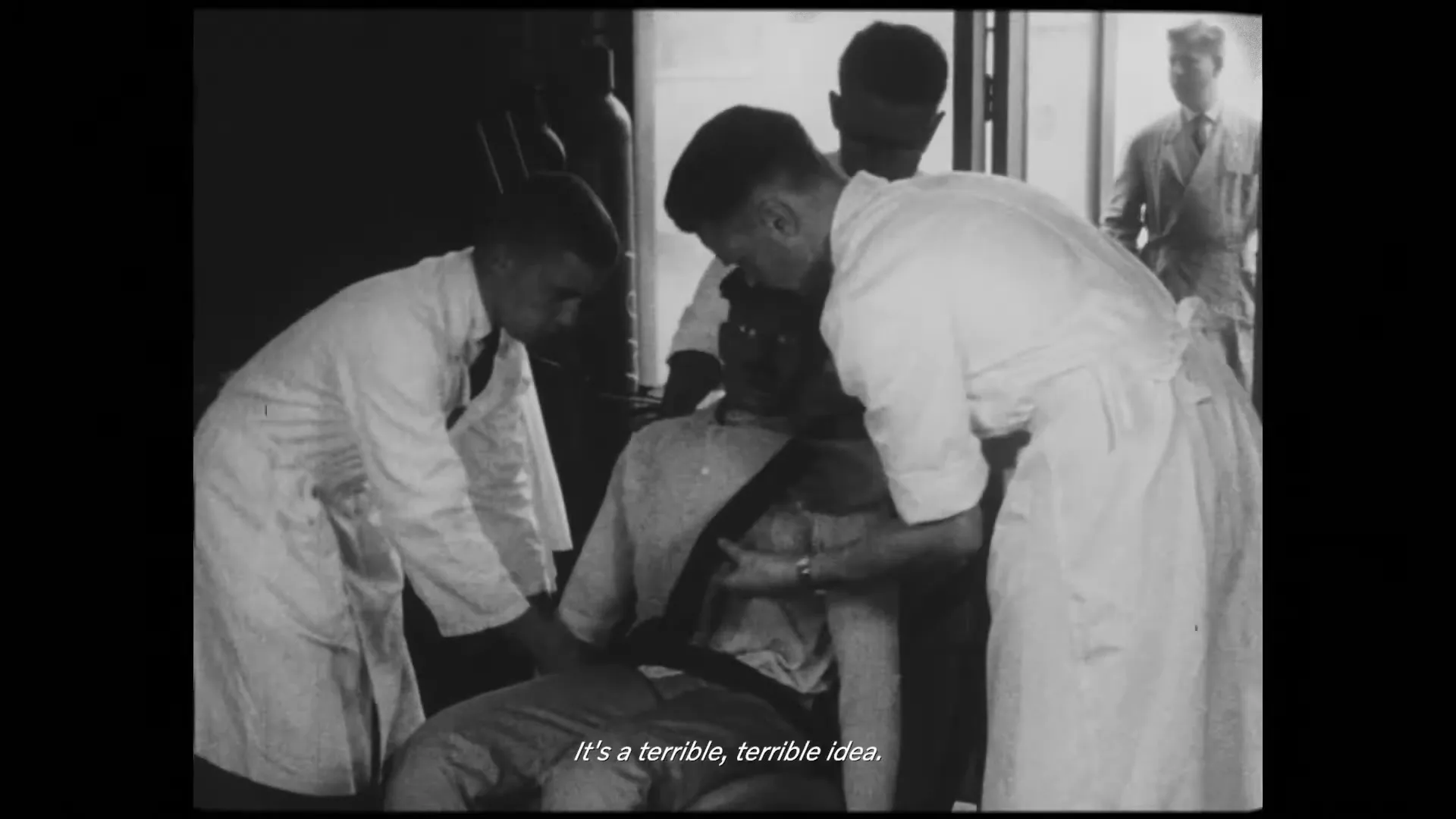In a black and white archival shot, a man in a cap is seated while three men in lab coats attend to him, seemingly performing a medical procedure.
