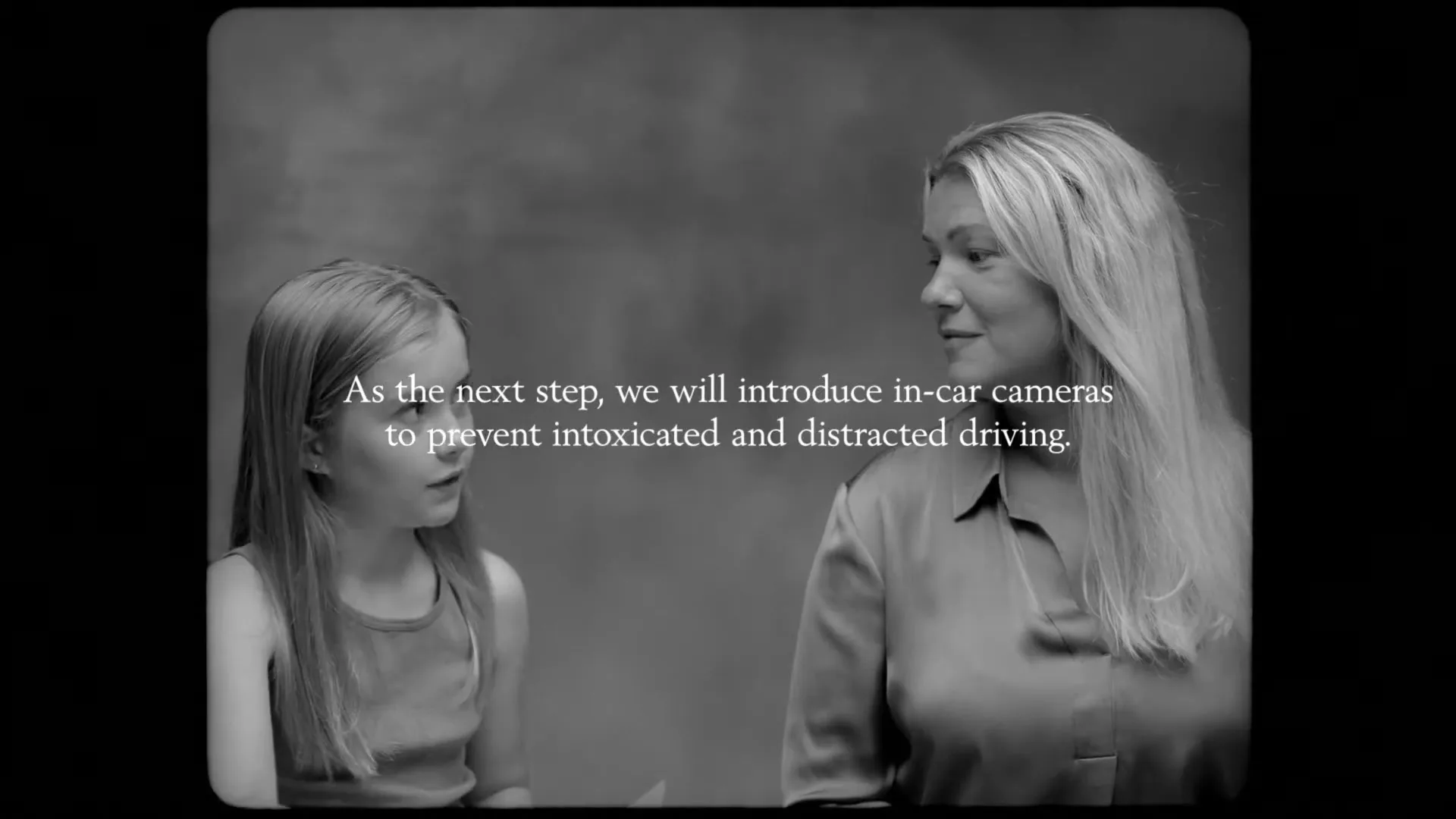 A young girl and a woman sit side-by-side, looking at each other and then at the camera, in a black and white studio setting.