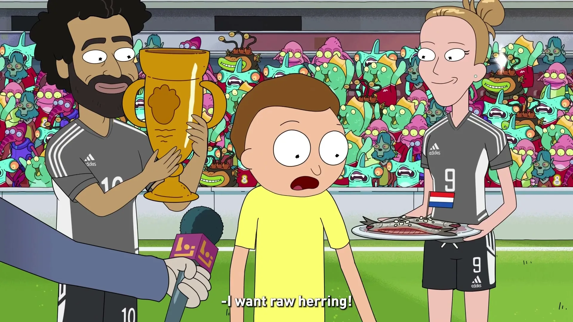 Morty Smith, a young boy in a yellow shirt, is interviewed on a soccer field, surrounded by alien fans, as two soccer players offer him a trophy and a plate of raw herring.