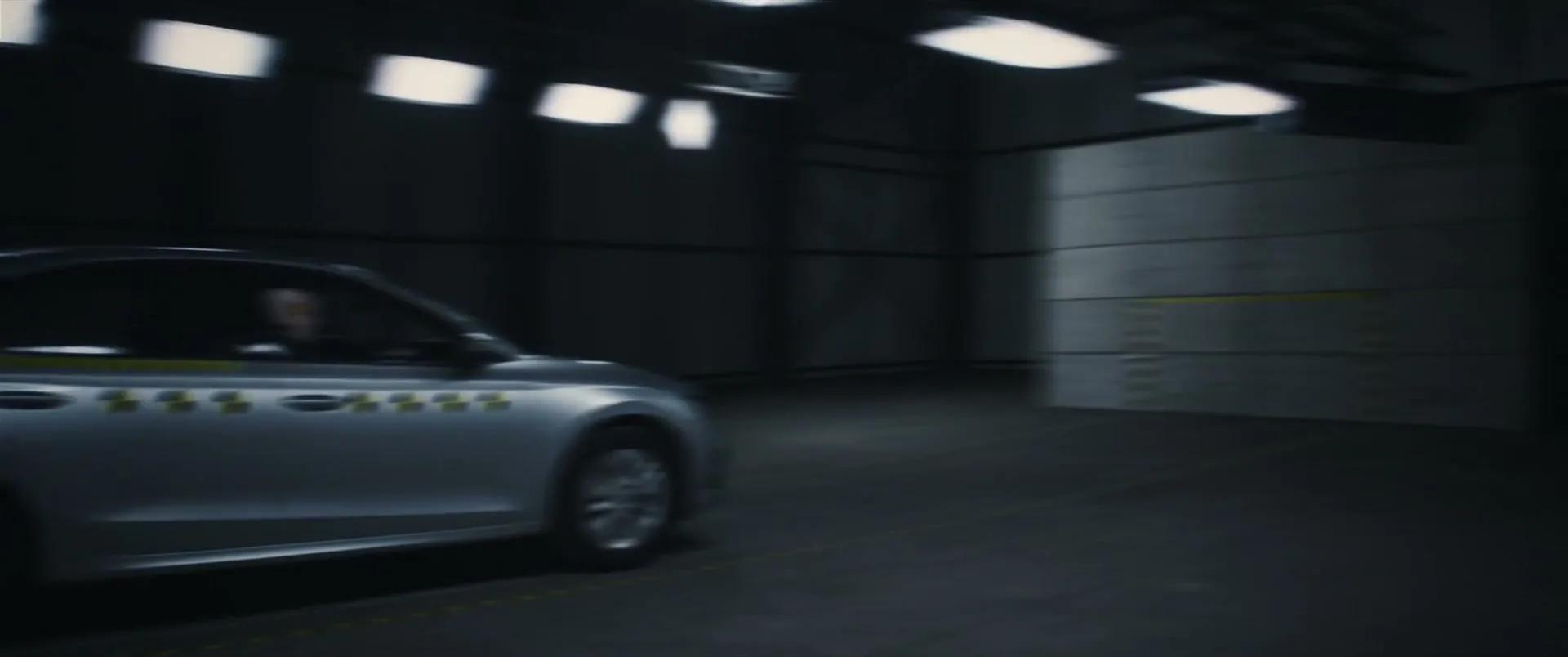 A crash test dummy in a silver car is shown in a dark crash test facility, then the scene transitions to a crash test dummy holding a baby dummy in a living room.