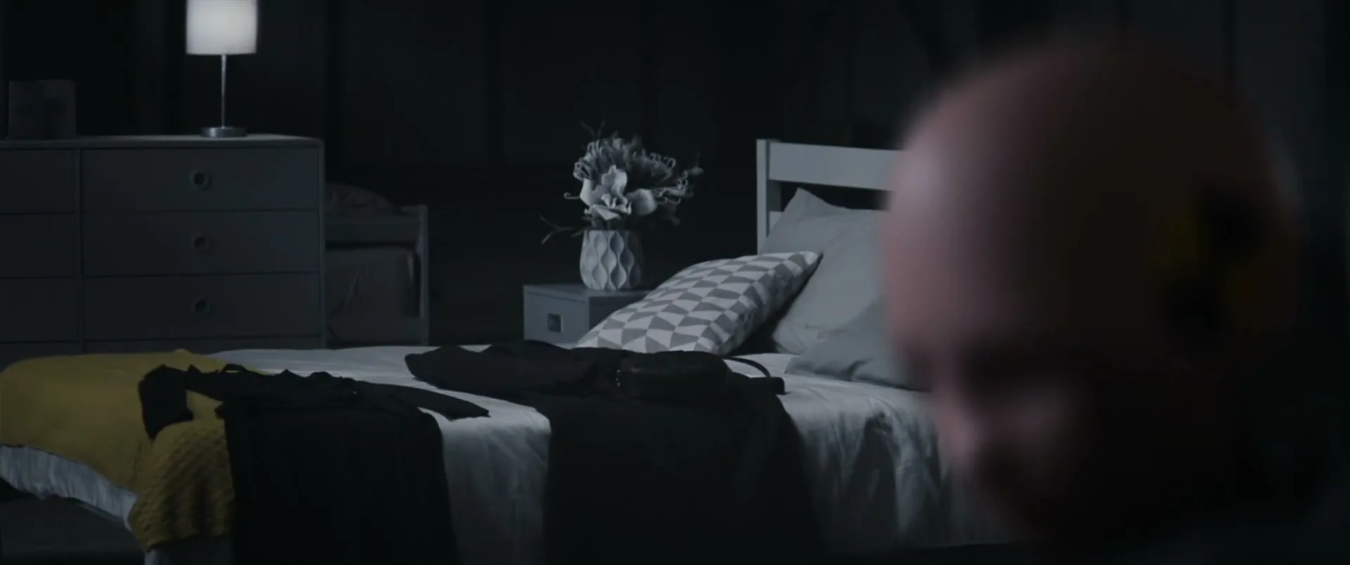 A crash test dummy's head is out of focus in the foreground, while a bedroom with clothes laid out on a bed is in focus in the background.