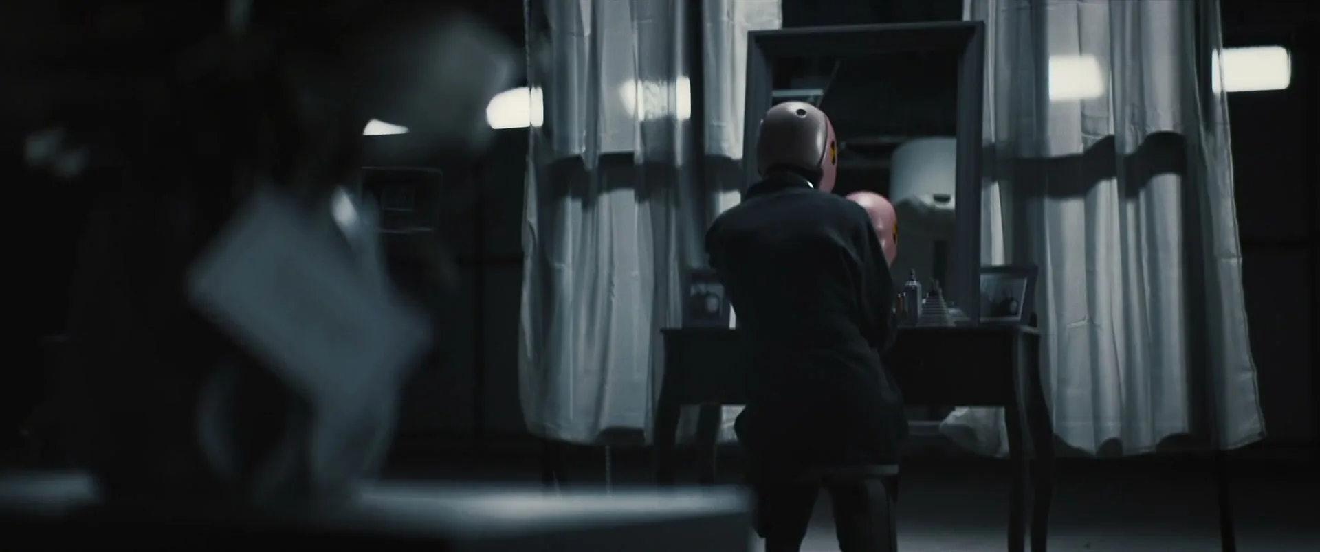 A crash test dummy in a suit sits at a vanity table, looking at its reflection in a mirror in a dark, industrial room.