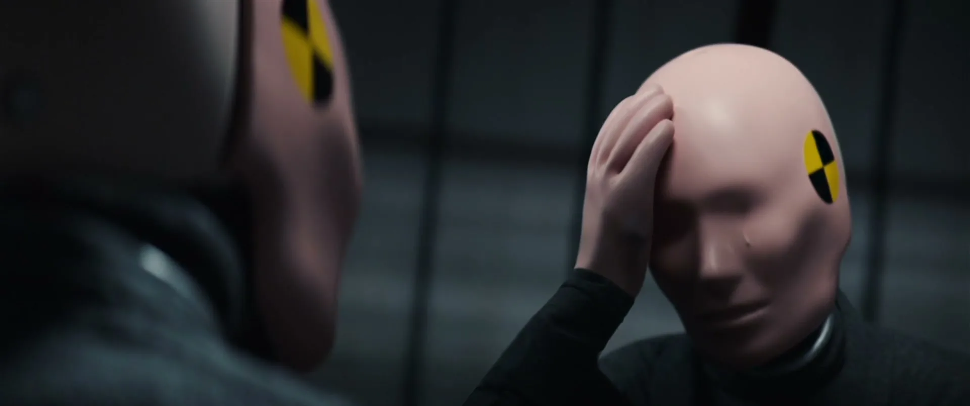 A crash test dummy in a dark suit holds its head in its hand, looking distressed, in a dark, industrial setting.