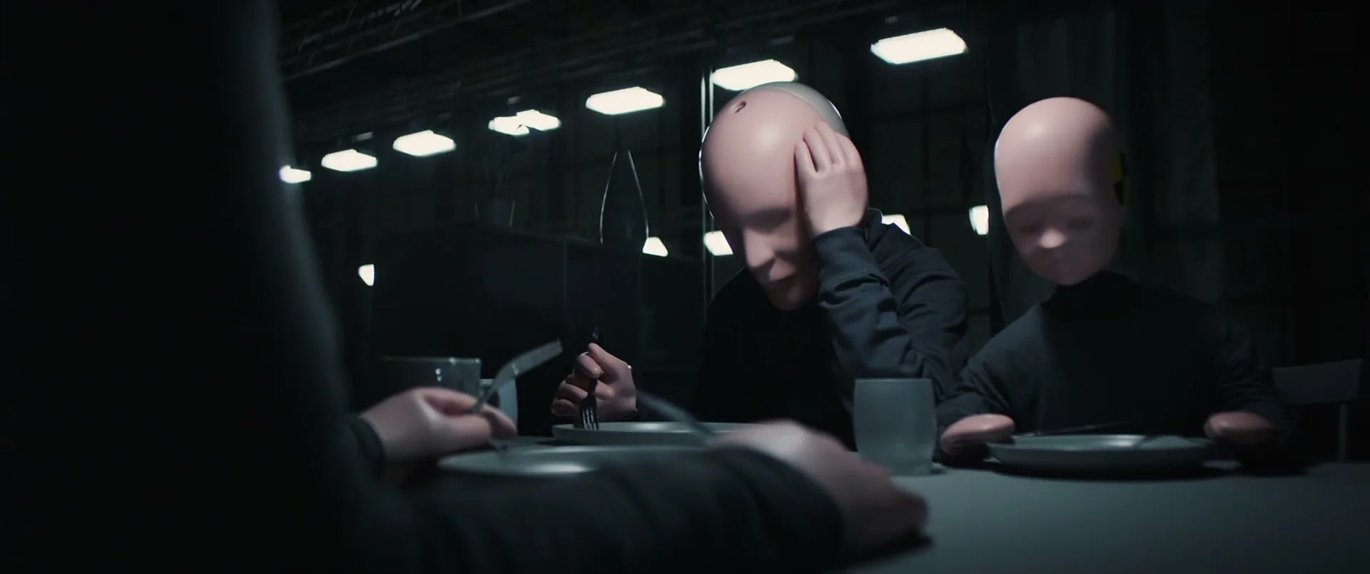 Three crash test dummies sit at a table in a dark, industrial setting, with one dummy resting its head in its hand.