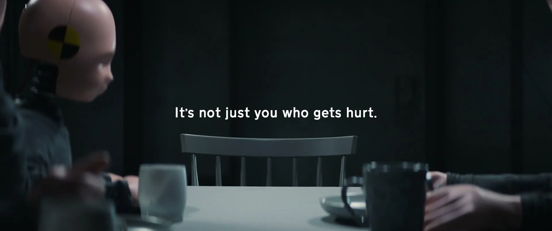Three crash test dummies, two adults and one child, sit around a table in a dark room, with text appearing on screen.