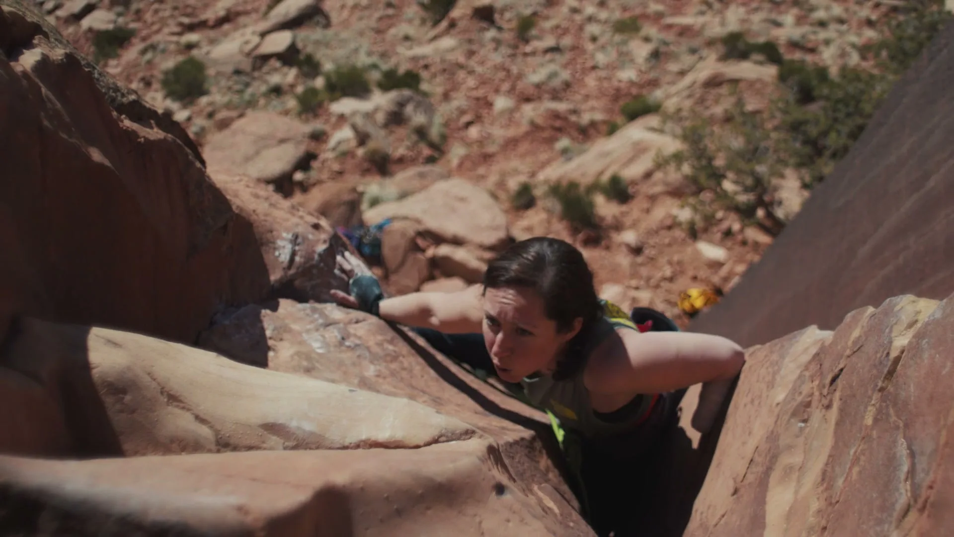 A female rock climber in a tank top and gloves struggles to ascend a narrow rock face in a desert canyon on a sunny day.
