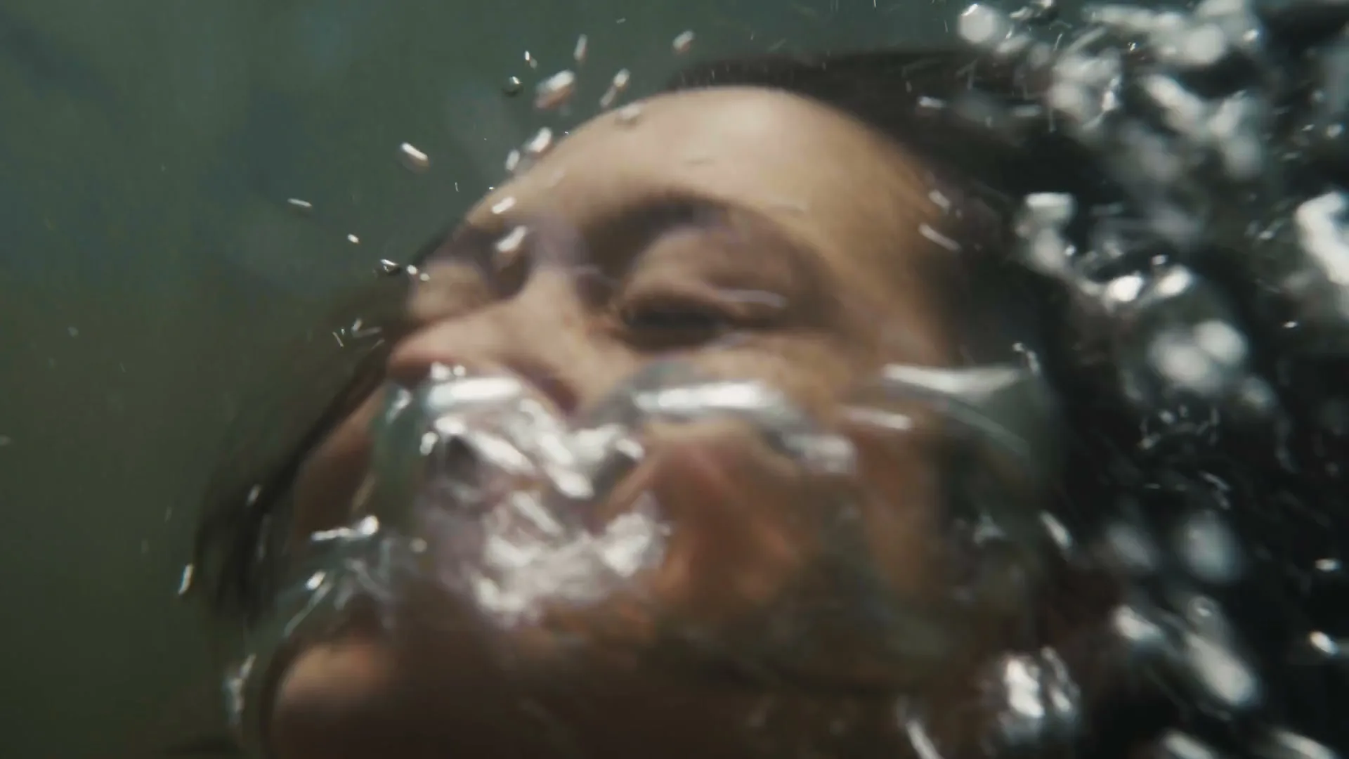 A woman with dark hair is seen underwater, surrounded by many bubbles, with her eyes closed and mouth slightly open.