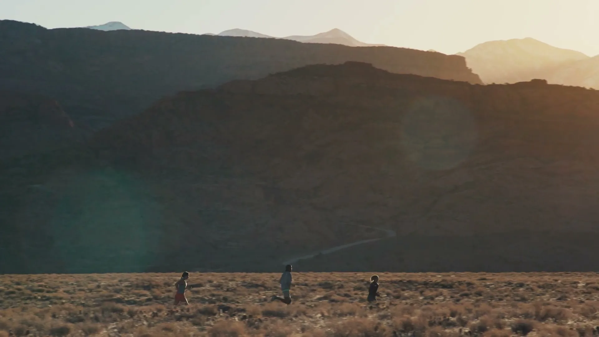 Three people run across a vast, dry field towards distant mountains under a bright sky, with a lens flare appearing.