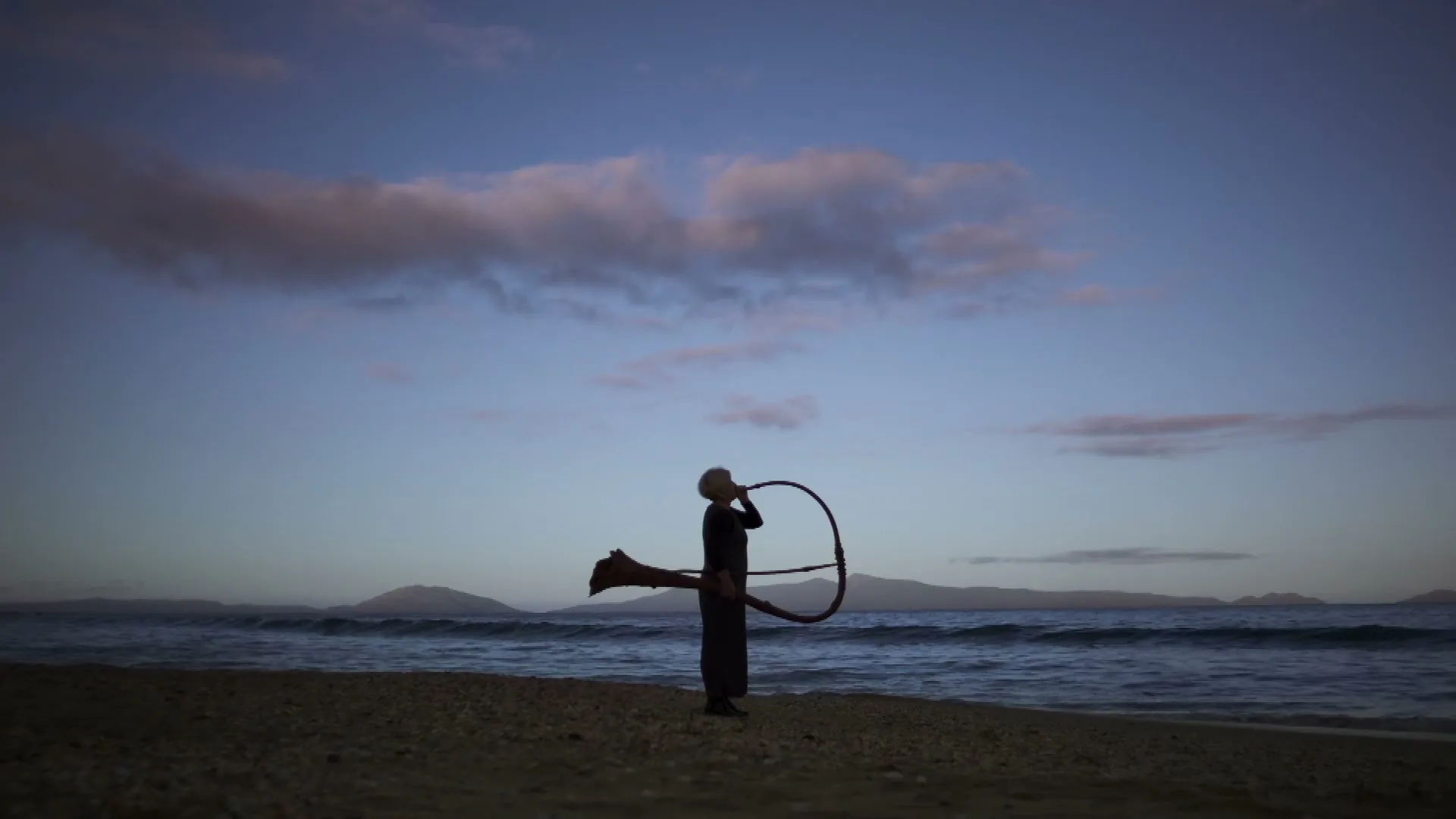 A lone figure in silhouette stands on a beach at dusk, playing a large, circular horn-like instrument.