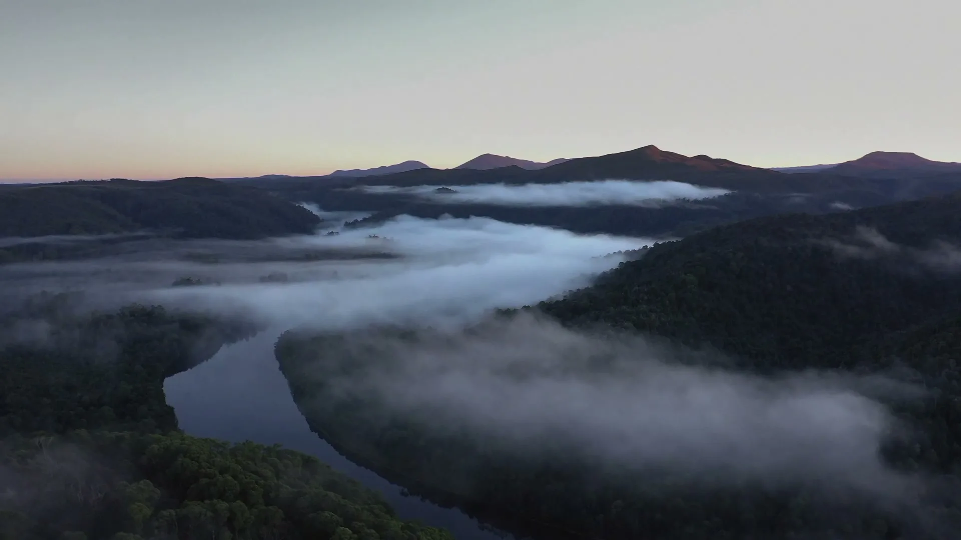 An aerial view of a winding river through a misty, forested valley with mountains in the background at dawn.