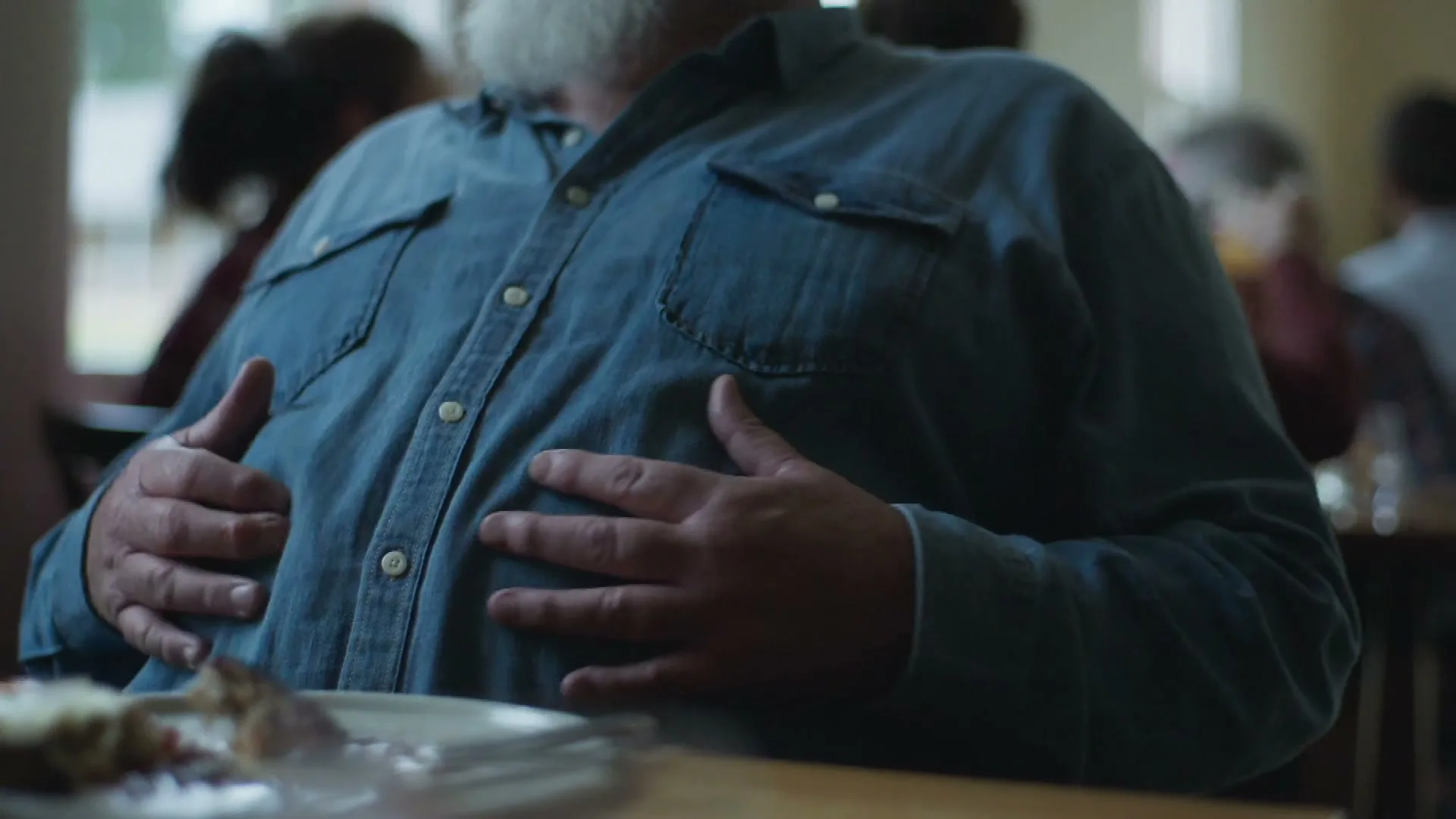 A man with a white beard, wearing a blue denim shirt, sits at a table in a restaurant, patting his full stomach after a meal.