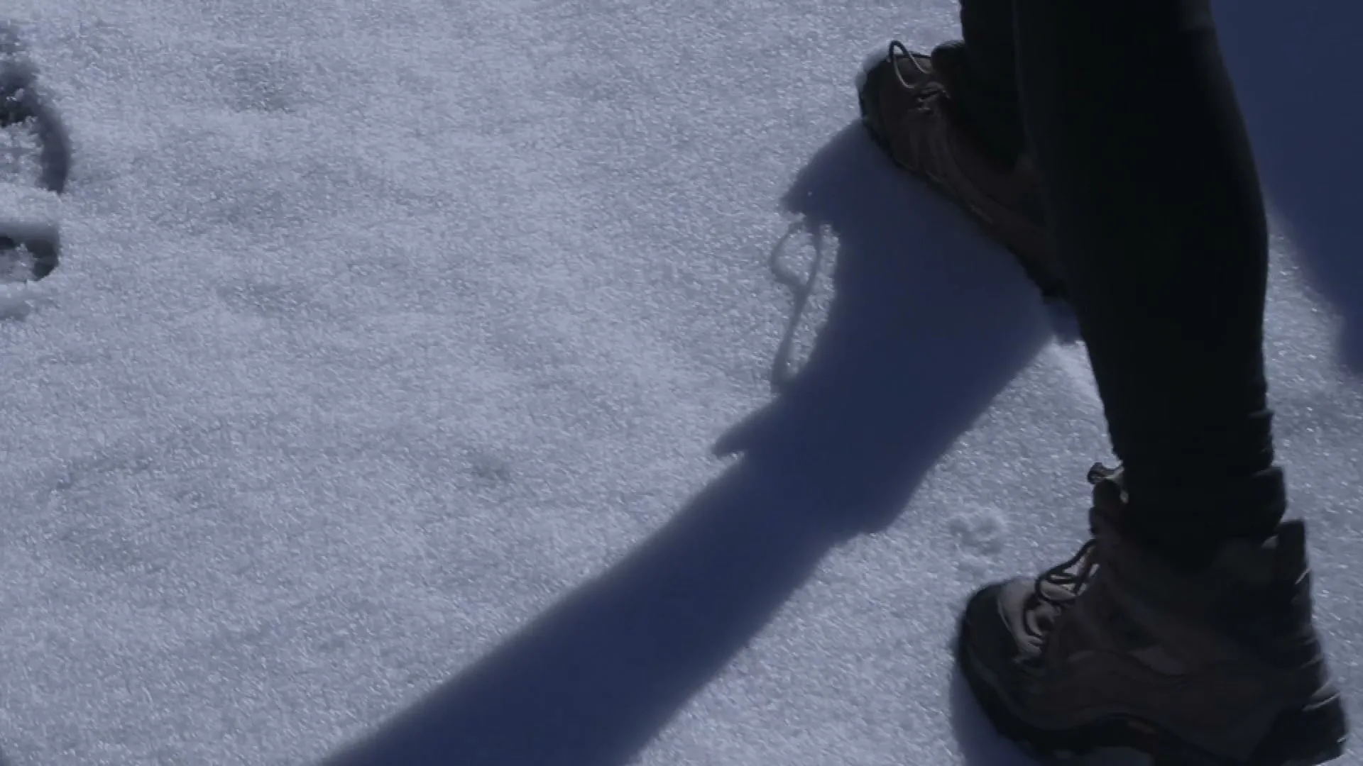 A person's legs and hiking boots are seen walking on a snowy surface, casting a long shadow in bright sunlight.