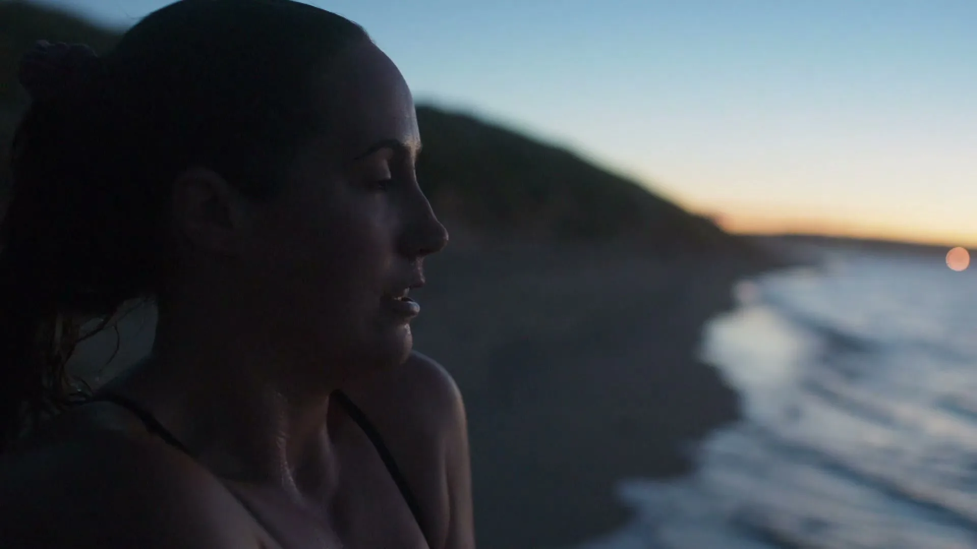 A woman with wet hair in a swimsuit looks out at the ocean at dusk, with a hill in the background.
