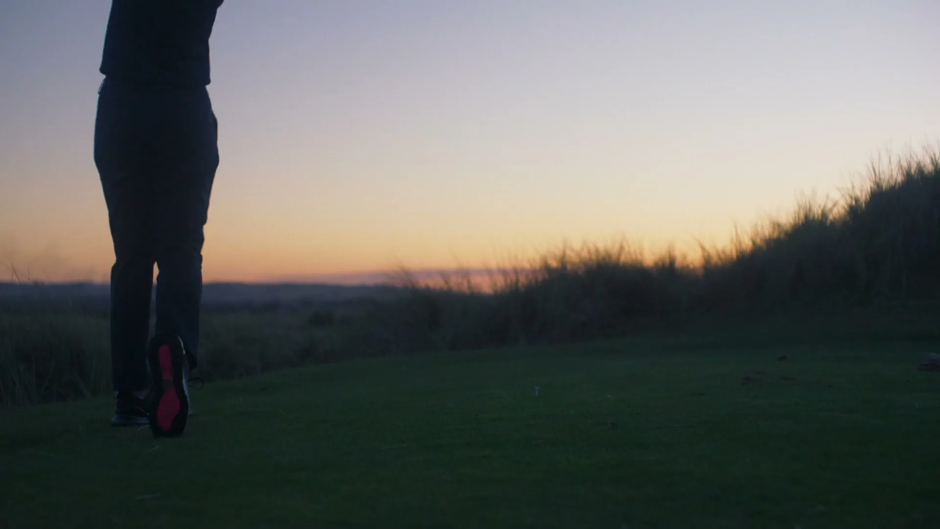 A person in dark athletic wear swings a golf club on a green course at dusk, with tall grass and a colorful sky in the background.