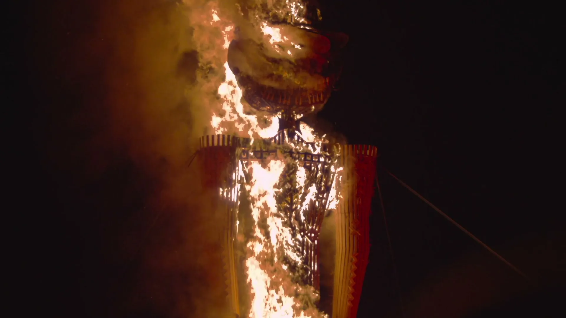 A large, human-shaped effigy made of wood and straw burns brightly against a dark night sky, with flames engulfing its body.