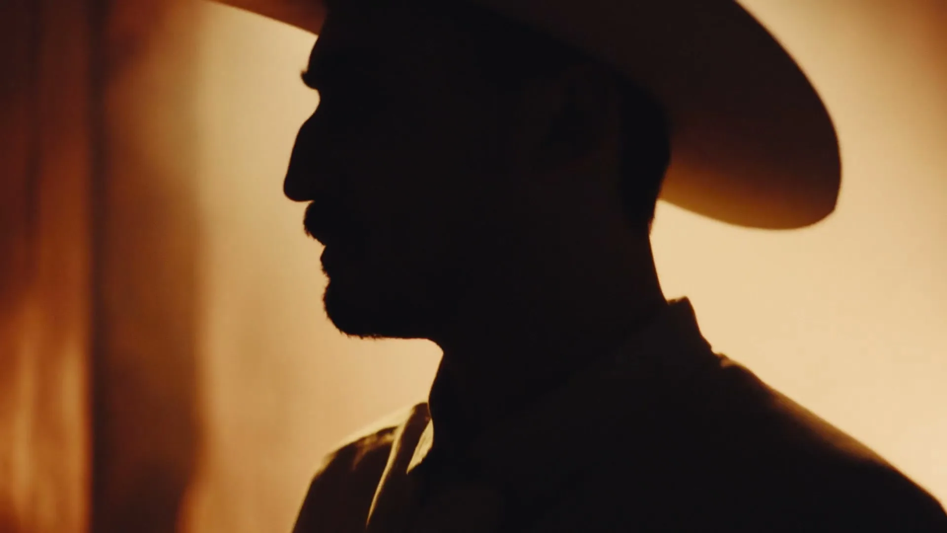 A man in a cowboy hat is silhouetted against a warm, glowing background, turning his head to the right.