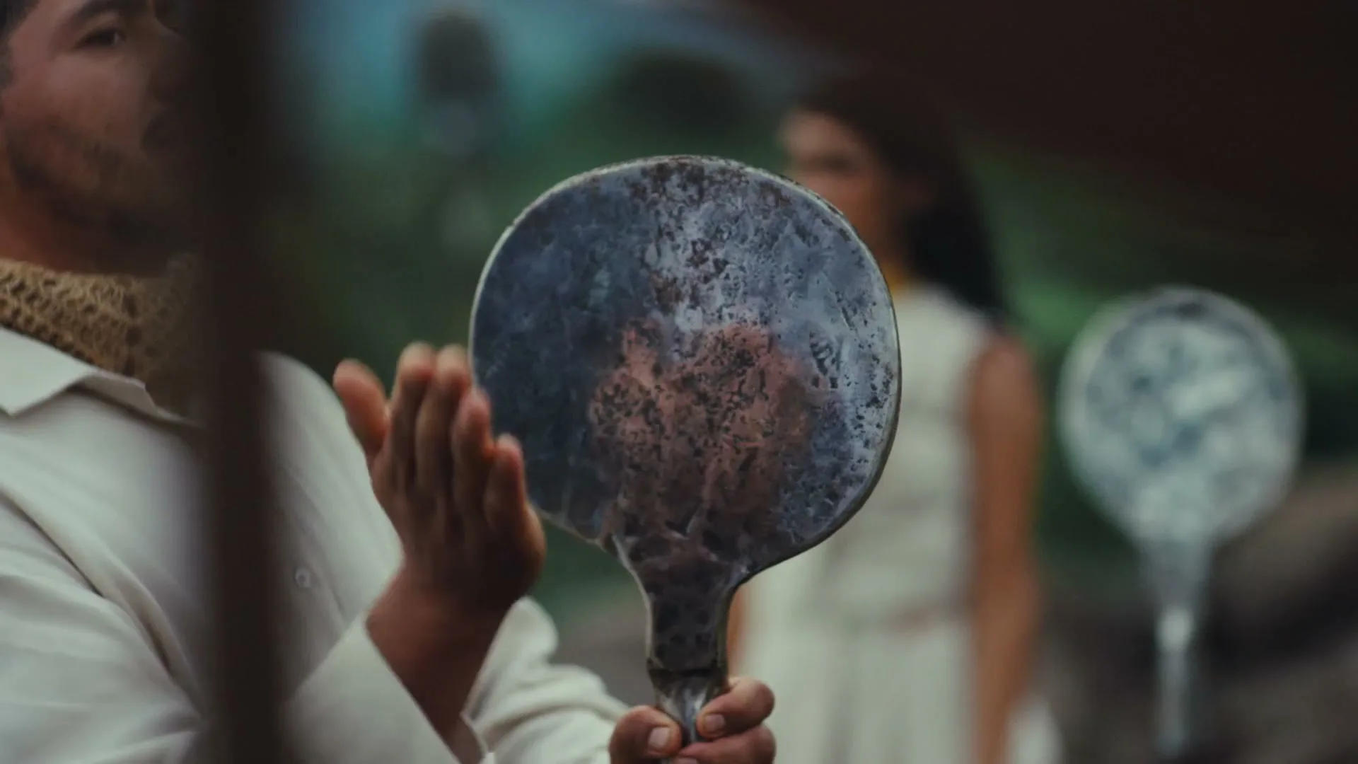 A man in a white shirt holds a metal disc, while a woman in a white dress stands in the background, outdoors.