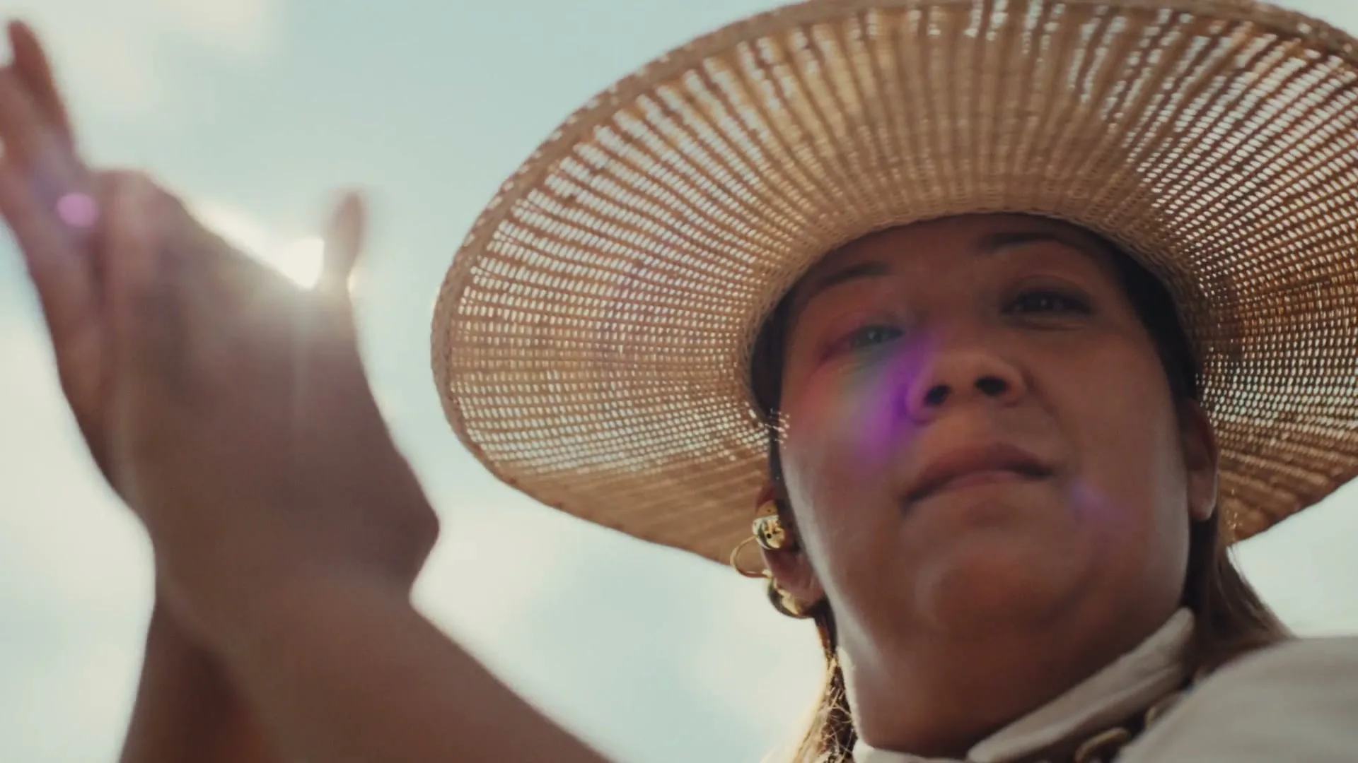 A woman in a straw hat claps her hands, looking up at the sky on a sunny day.