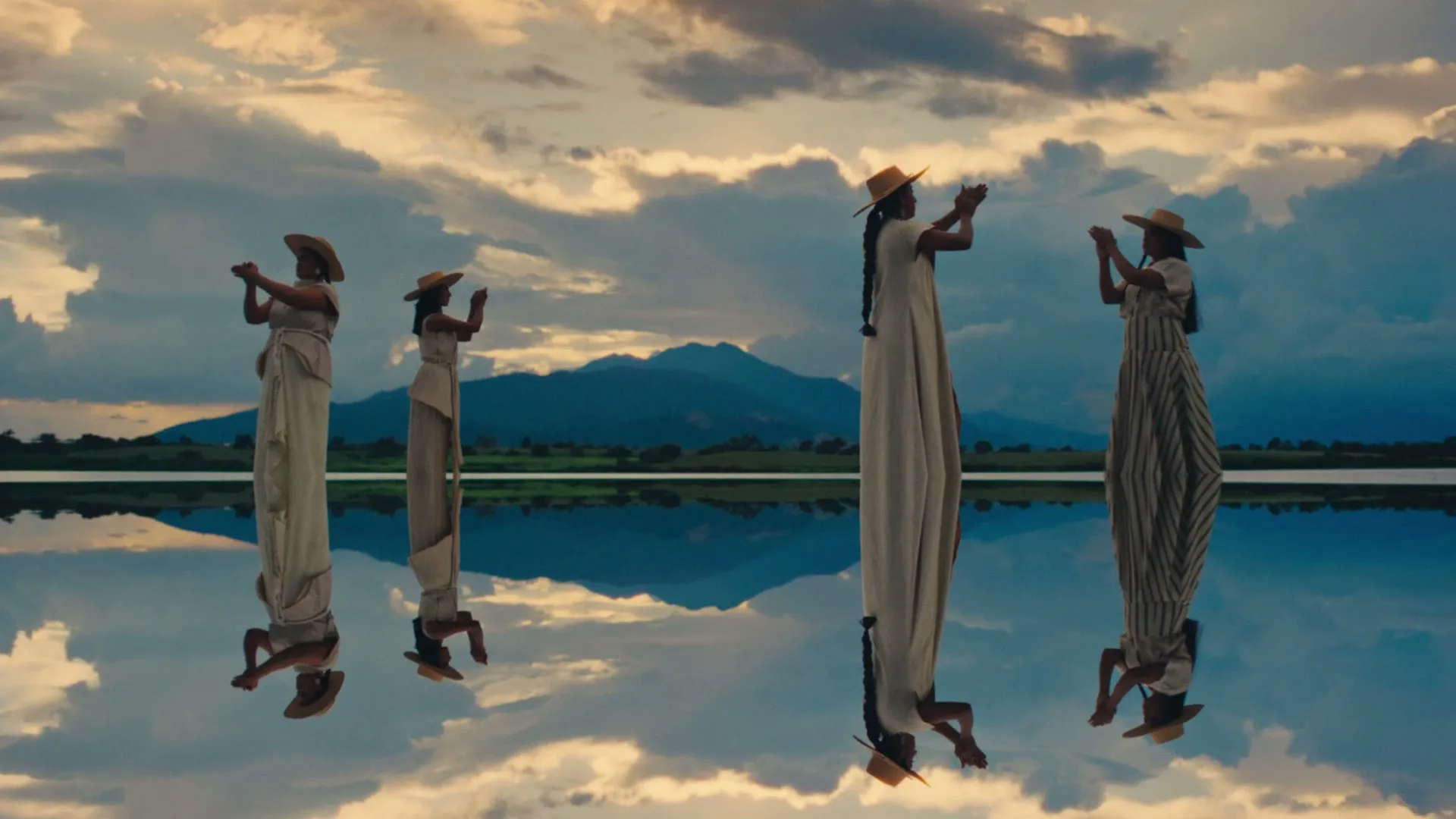 Four women in long dresses and straw hats stand on a reflective surface, with mountains and a cloudy sky in the background, creating a mirrored effect.