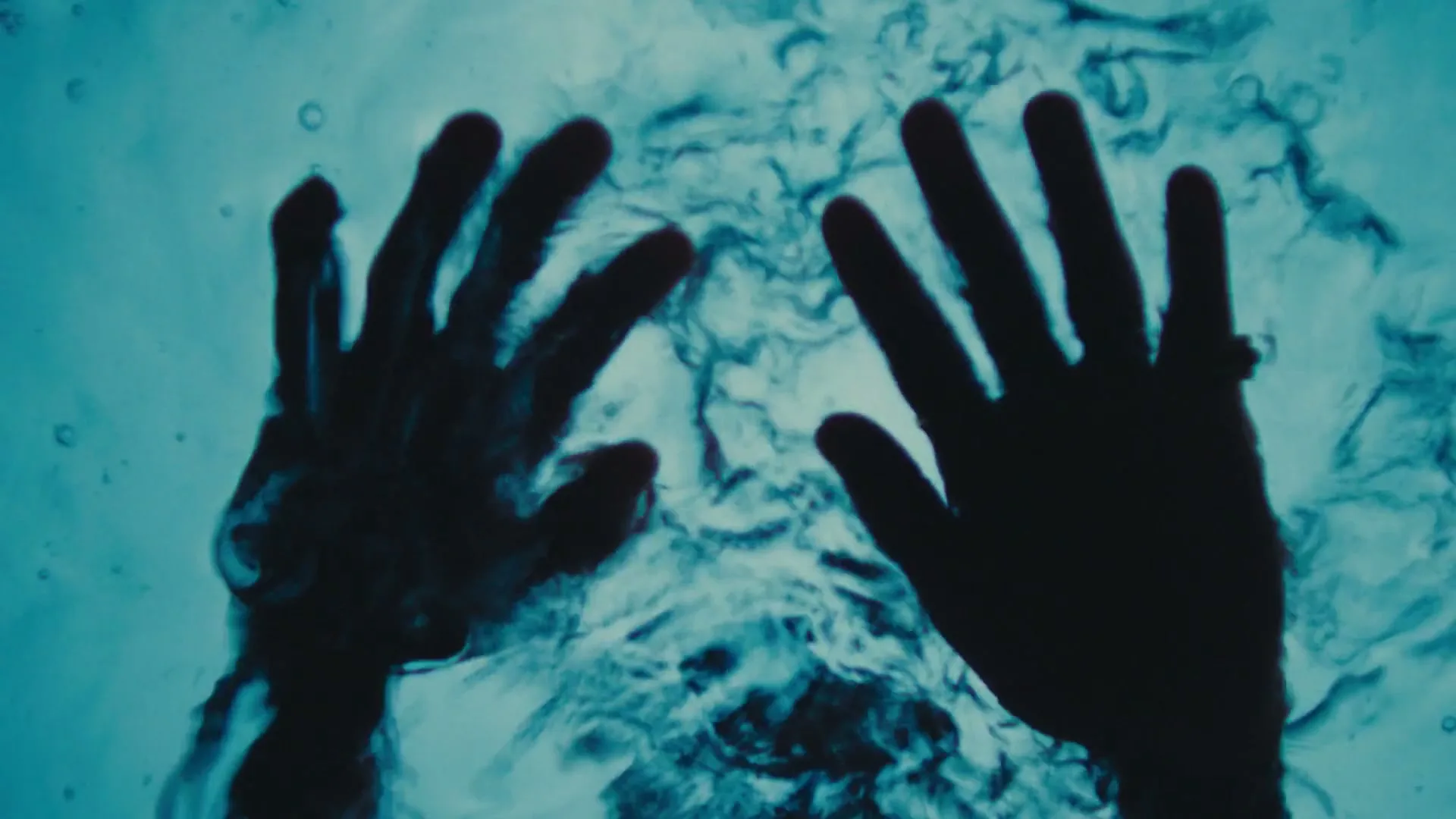 Two silhouetted hands are seen from below, pressing against a rippling, turquoise water surface, creating dynamic patterns.