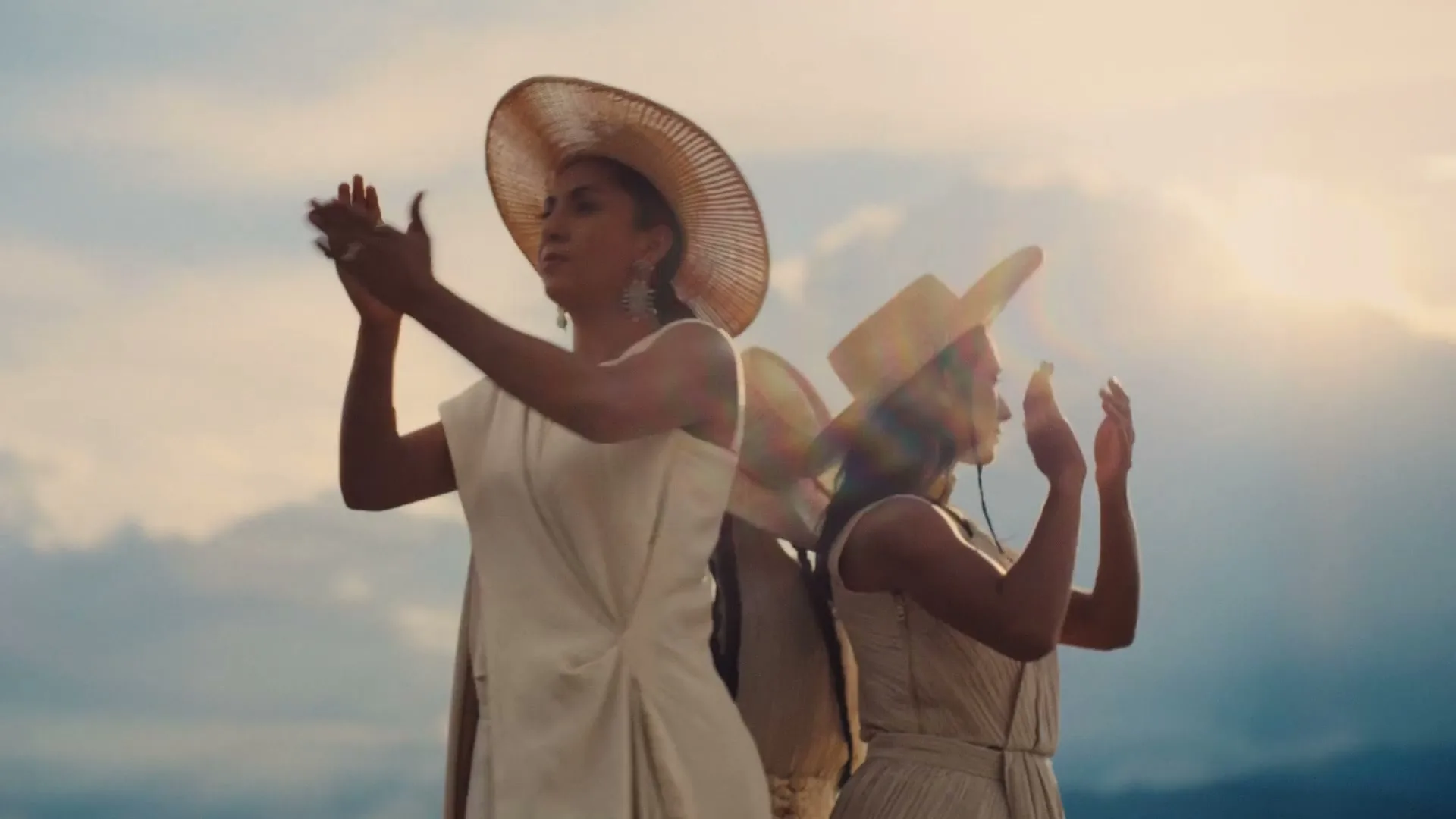 Two women in straw hats and light dresses stand with their hands raised, silhouetted against a cloudy sky at golden hour.