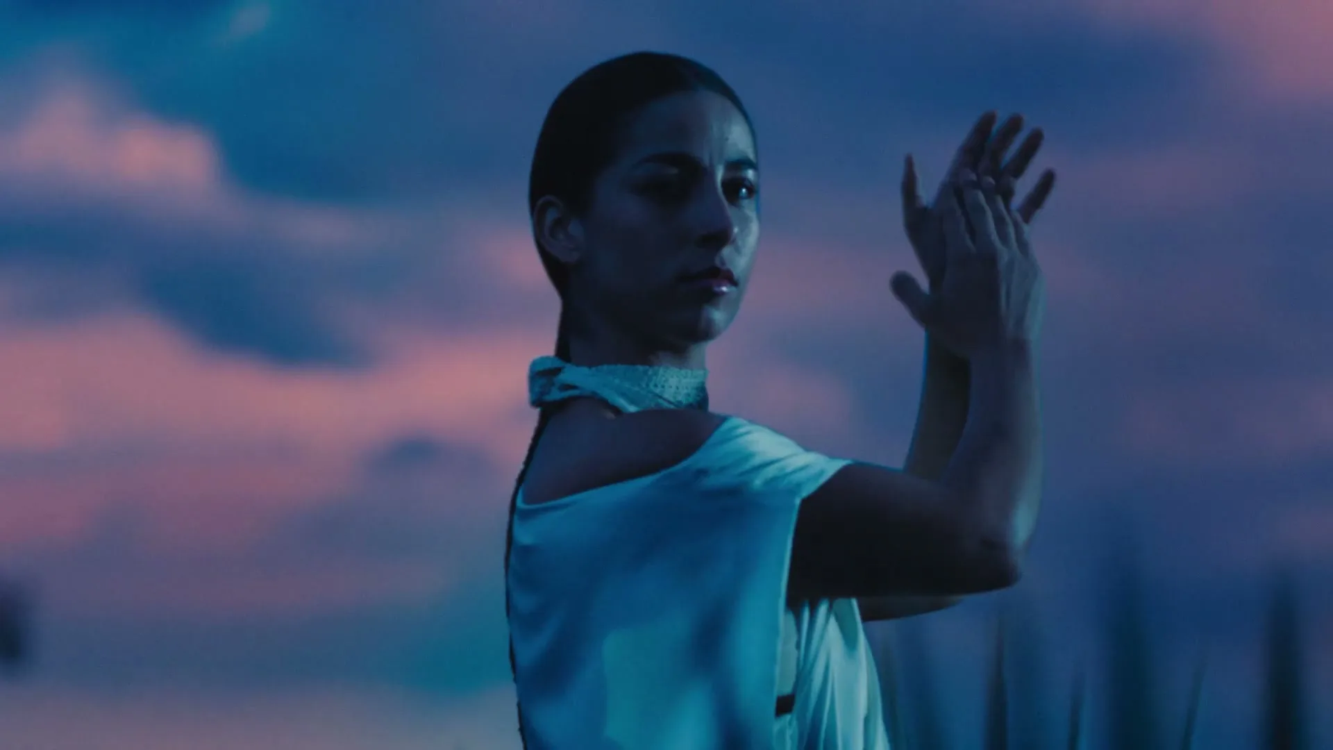 A woman with dark hair in a white dress claps her hands, looking intently at something off-screen, against a dramatic blue and pink sky.