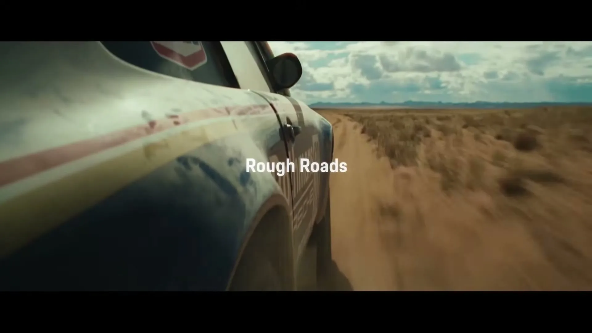 A dirty, white and blue Porsche 911 Dakar with racing stripes drives fast on a dusty, rough road in a desert landscape under a cloudy sky.