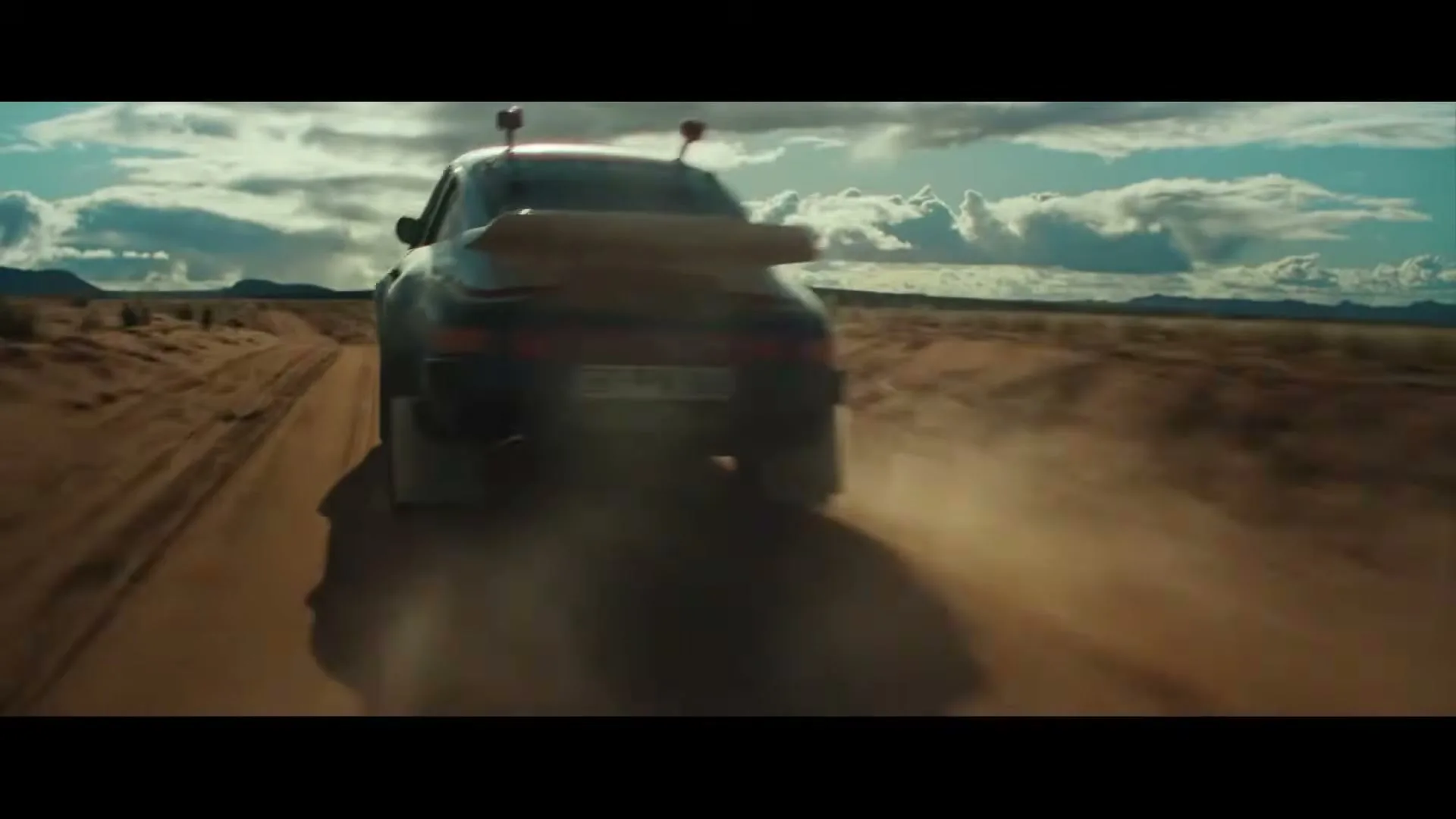 A dark Porsche 911 Dakar drives away from the camera on a dirt road, kicking up dust in a vast desert landscape under a cloudy sky.