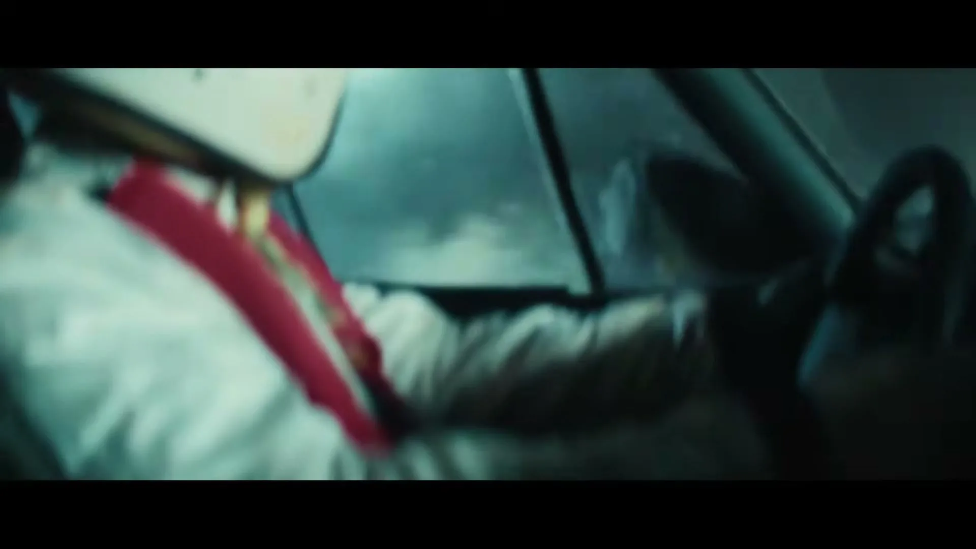 A blurry shot of a person in a white racing suit and helmet, with red straps, driving a car in what appears to be a dark, rainy environment.